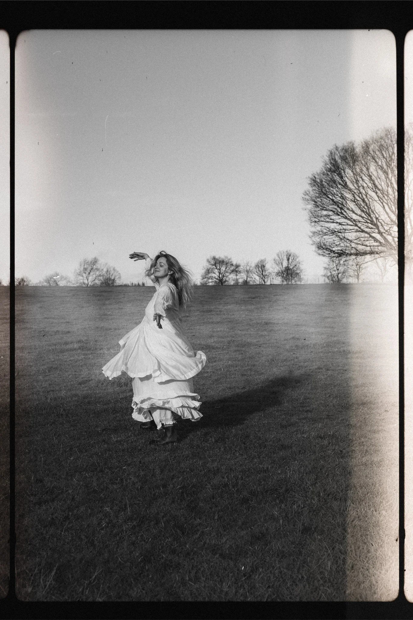 A woman dancing in a flowing dress on an open grassy field with trees in the background, captured in black and white.
