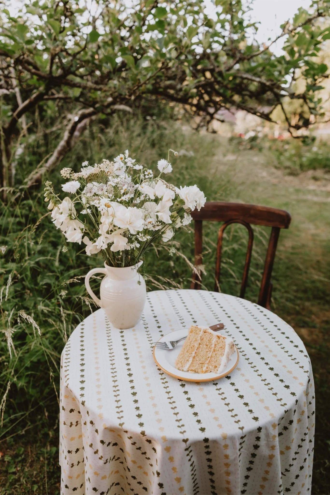 A round table with a patterned tablecloth outdoors, featuring a white vase with a bouquet of white flowers, a plate with a slice of cake, and a fork, with a wooden chair and greenery in the background.