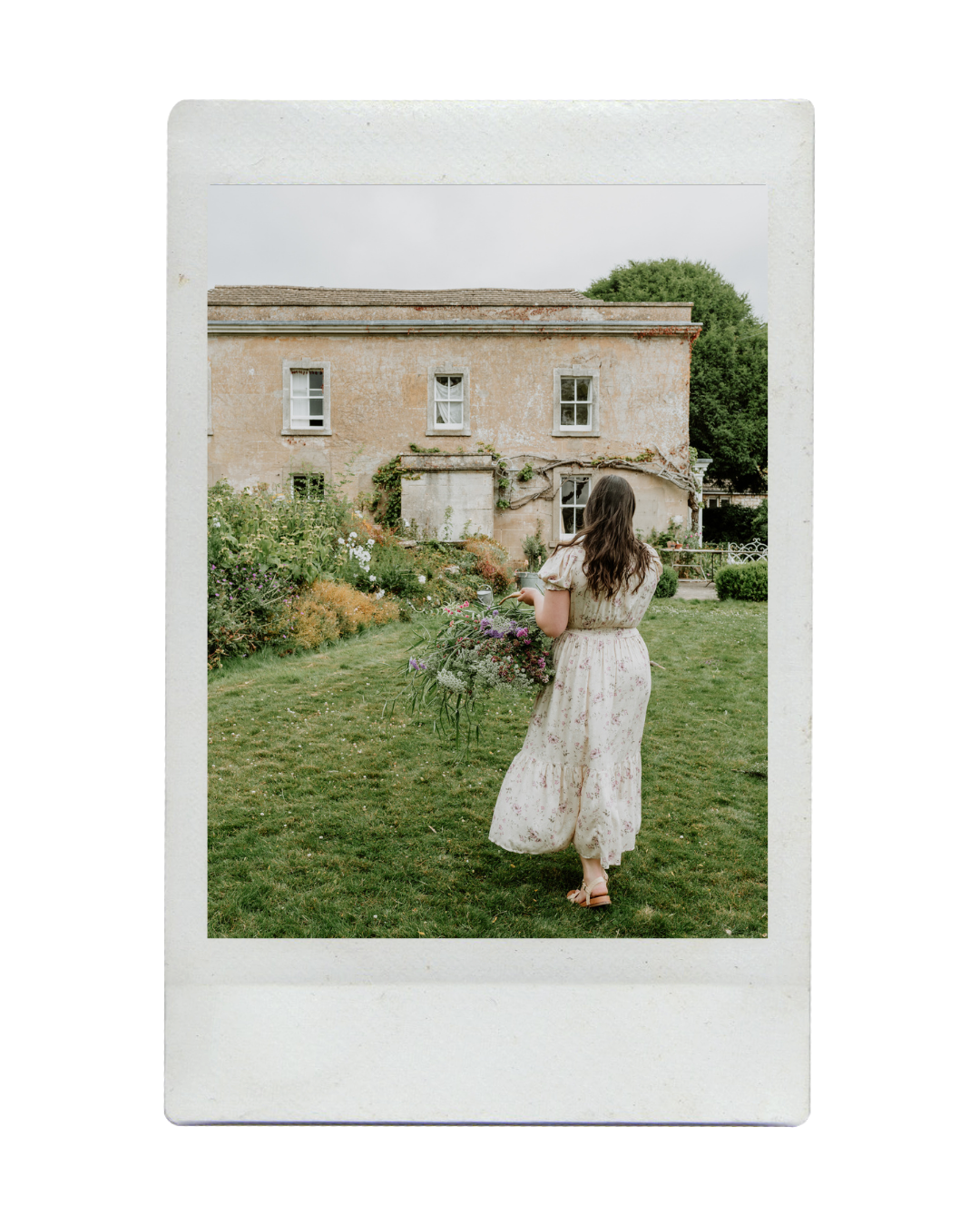 A woman in a vintage floral dress walking in a garden holding a large bouquet of flowers with an old rustic house in the background.