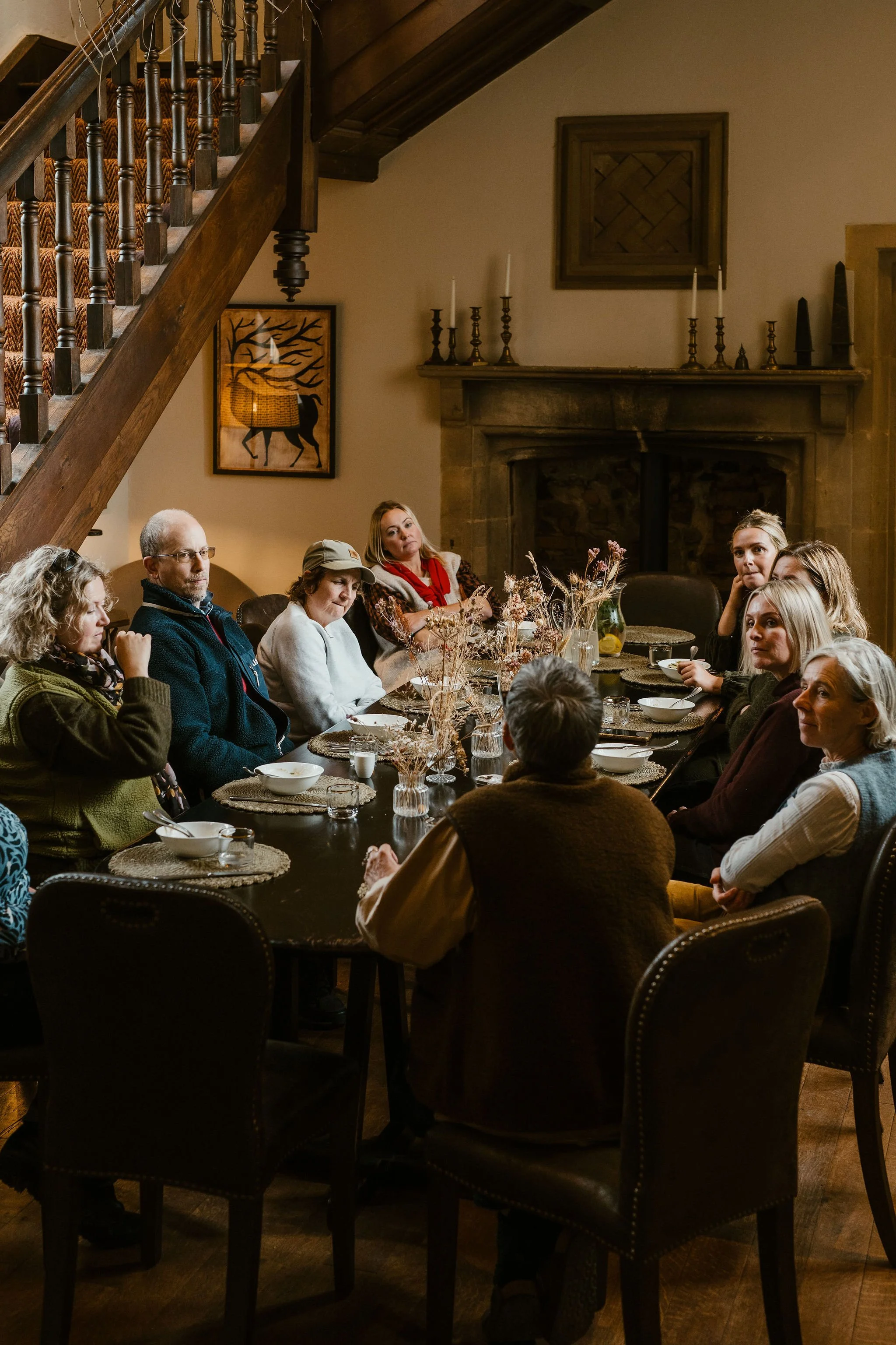 A group of people sitting around a large dining table in a cozy, warmly lit room with a stone fireplace and wooden staircase.