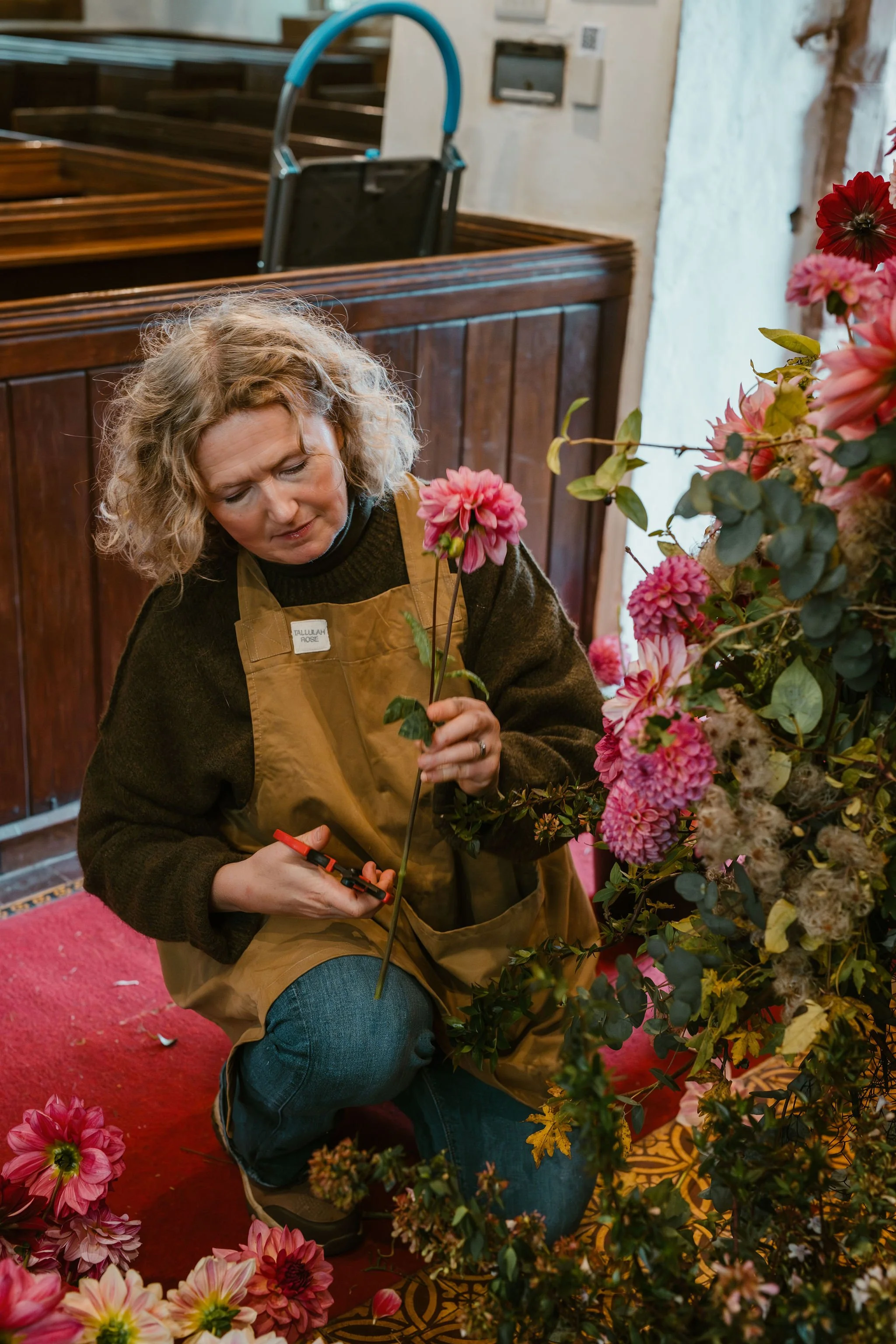 A woman kneeling and working with pink and white flowers in a flower shop, wearing a brown apron and holding a pair of pruning shears.