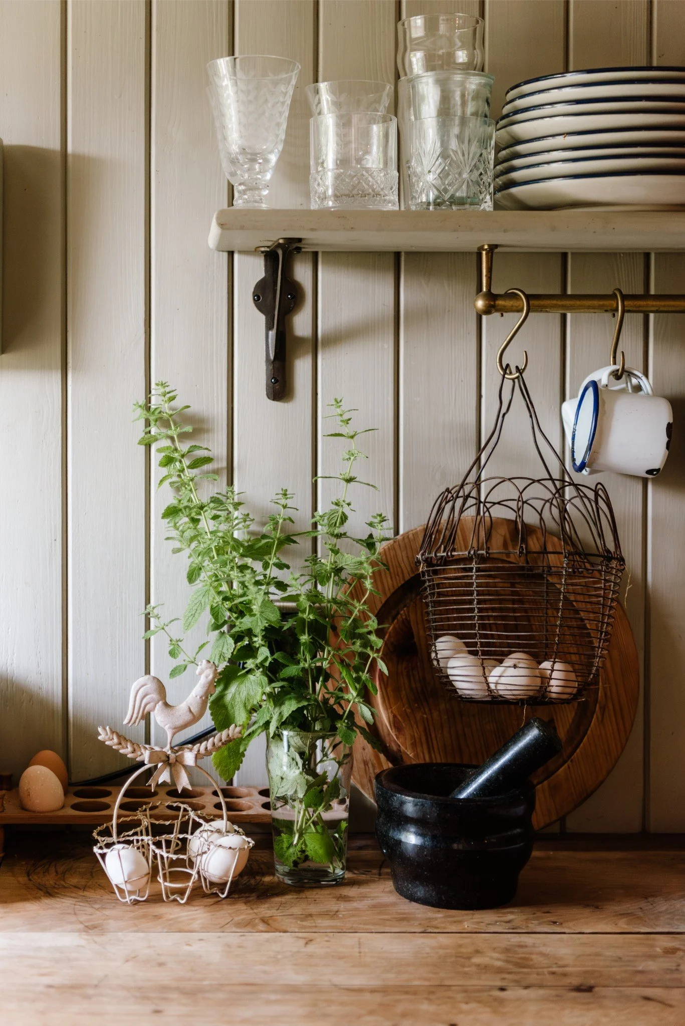 Kitchen scene with a wooden countertop, glass vase with green herbs, black mortar and pestle, hanging wire basket with eggs, wooden cutting board, plates, glasses, and cups on shelves.