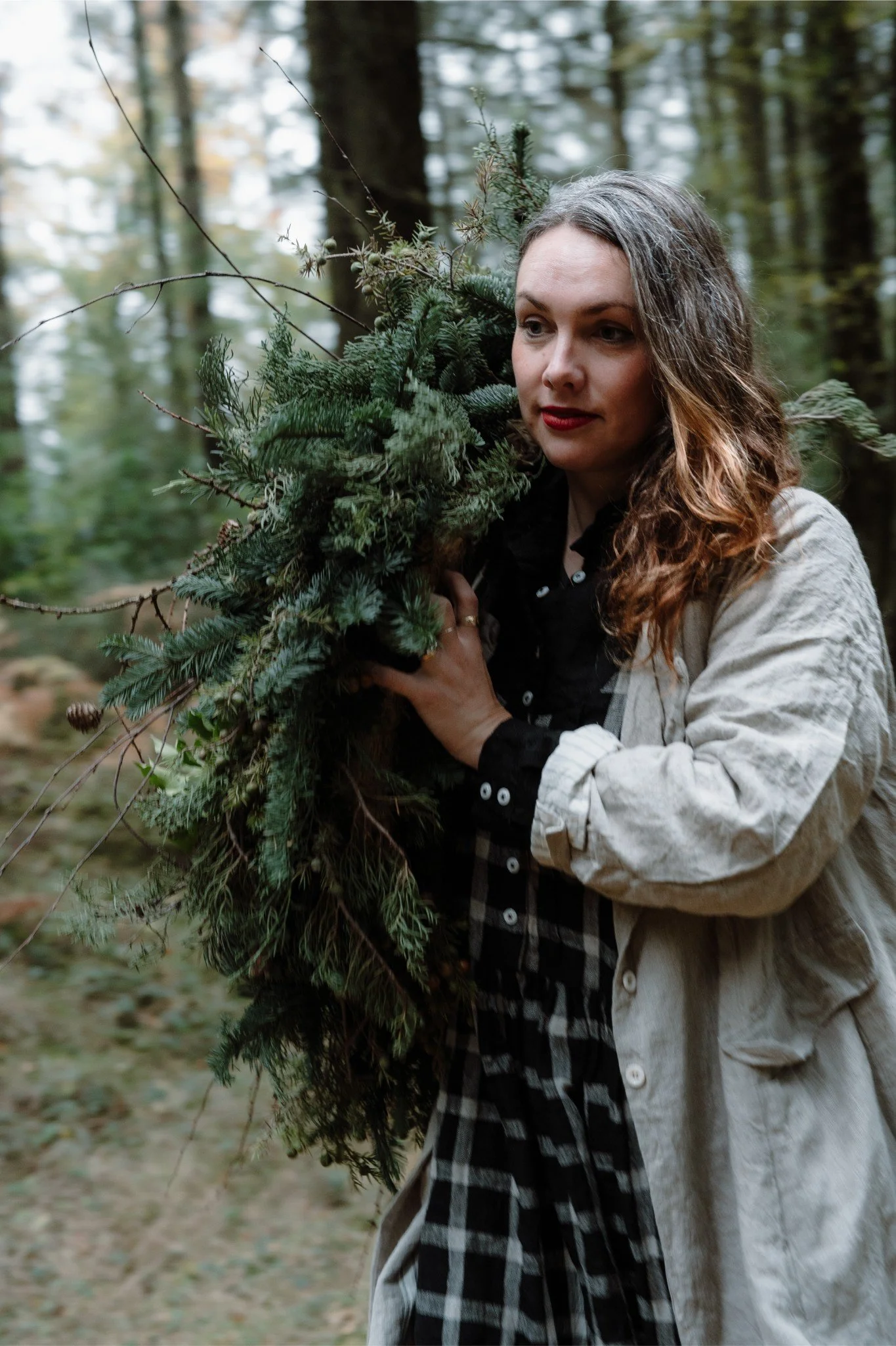 A woman carrying a large bundle of evergreen branches on her shoulder in a forest setting.