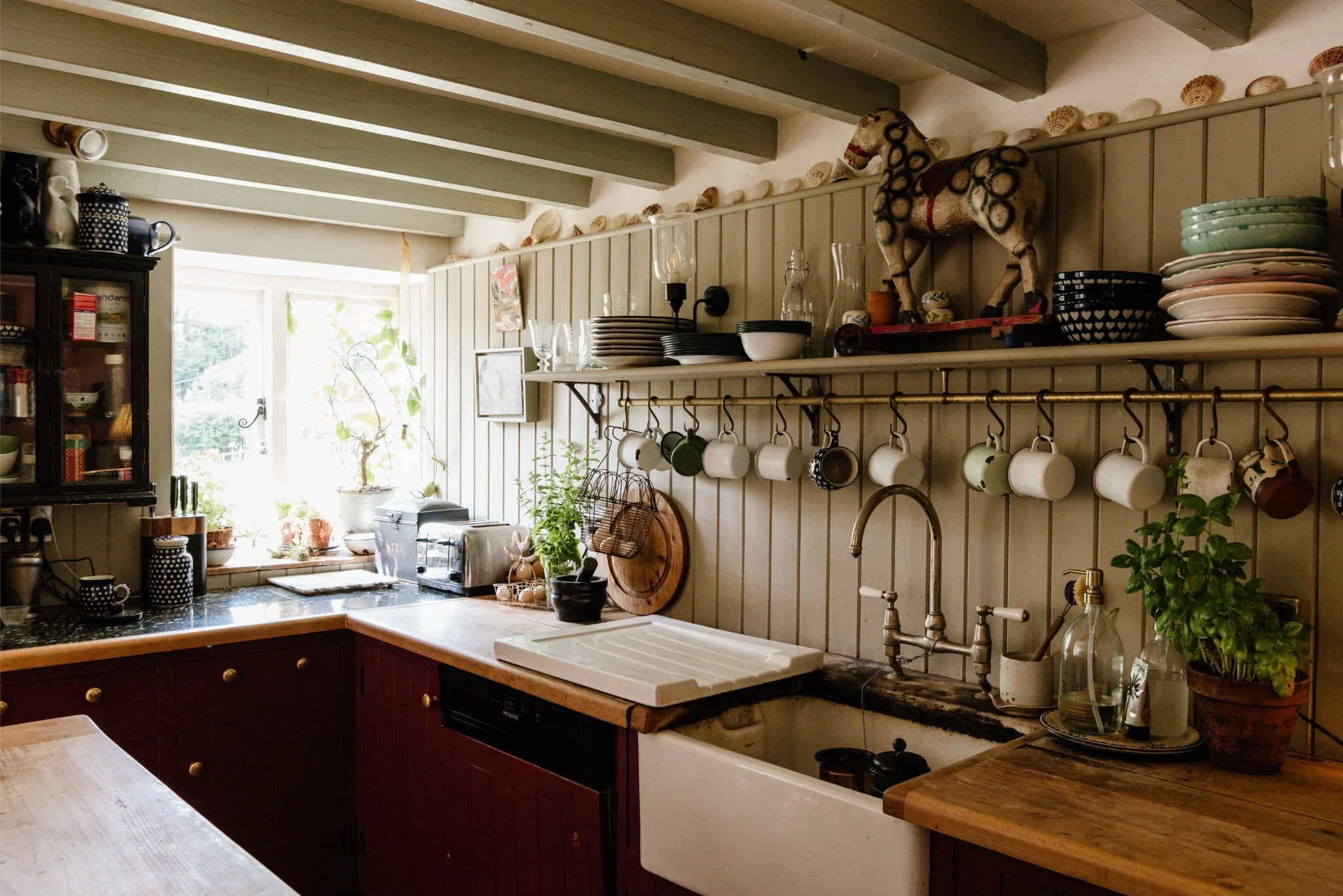 A cozy kitchen with a window letting in natural light, displaying open shelves with plates, bowls, and a decorative horse figure, hanging mugs, a sink with a rustic countertop, potted plants, and various kitchen utensils and appliances.