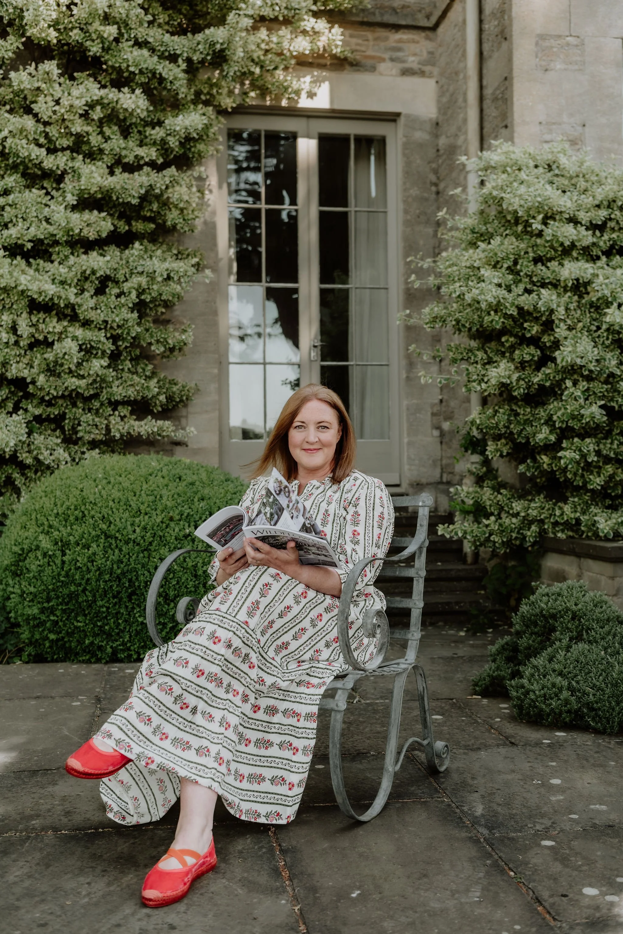 A woman with reddish hair sitting on a decorative metal bench outdoors, holding a magazine, wearing a long patterned dress and red shoes, with greenery and a stone building in the background.