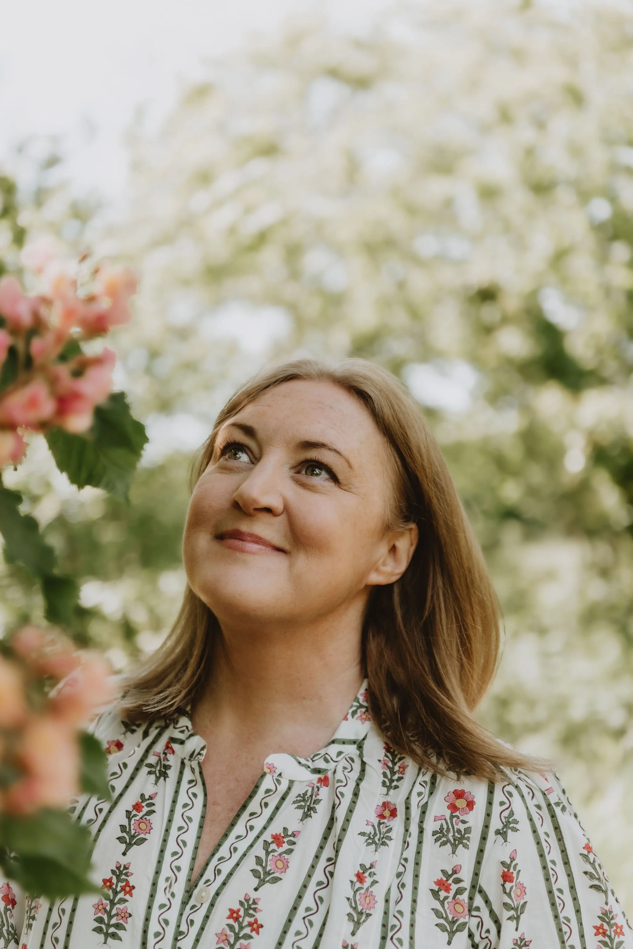 A woman with shoulder-length brown hair smiling while looking up at pink flowers on a tree in a garden or park.