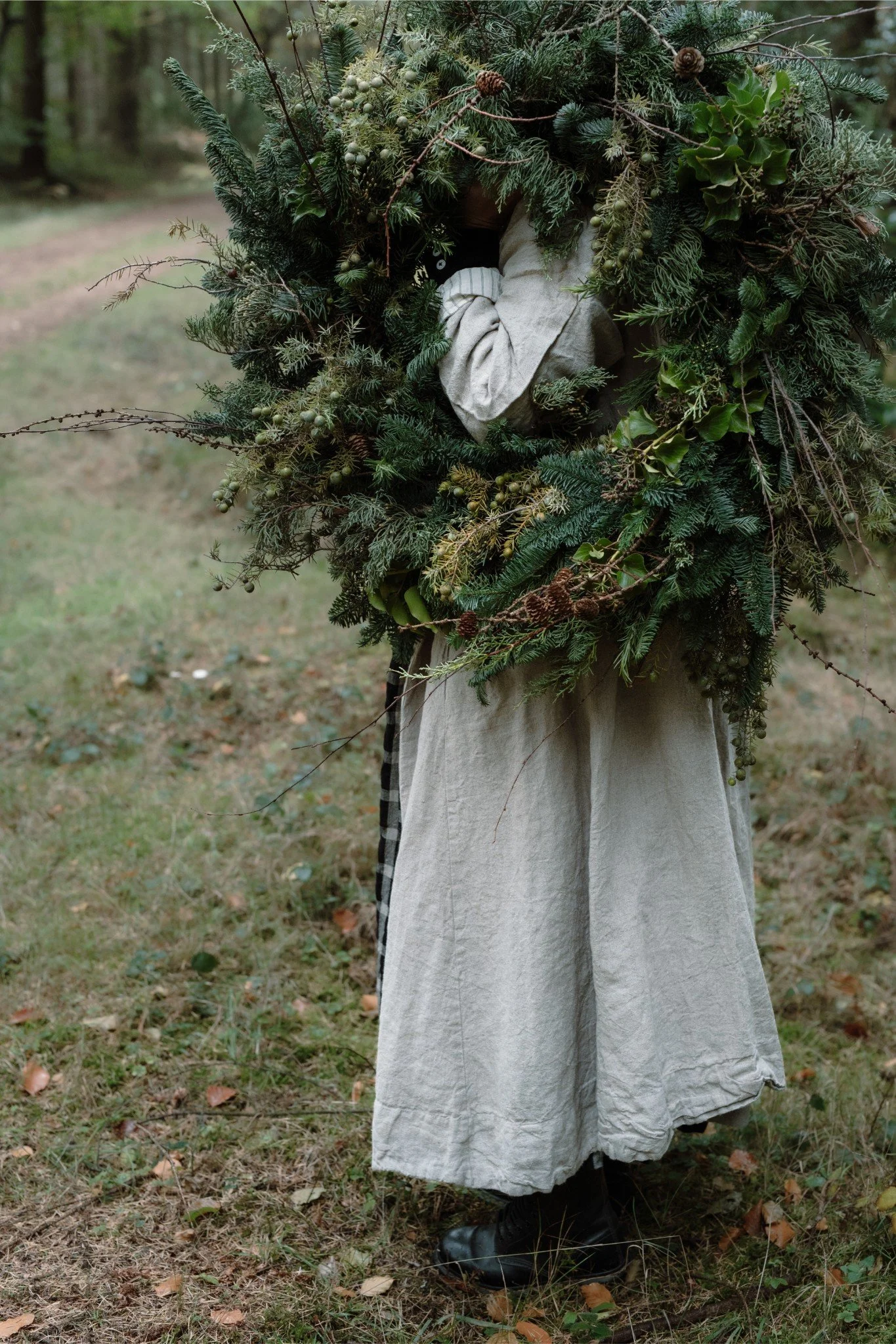 Person holding a large, lush Christmas wreath made of greenery and pinecones, standing outdoors on a grassy area.