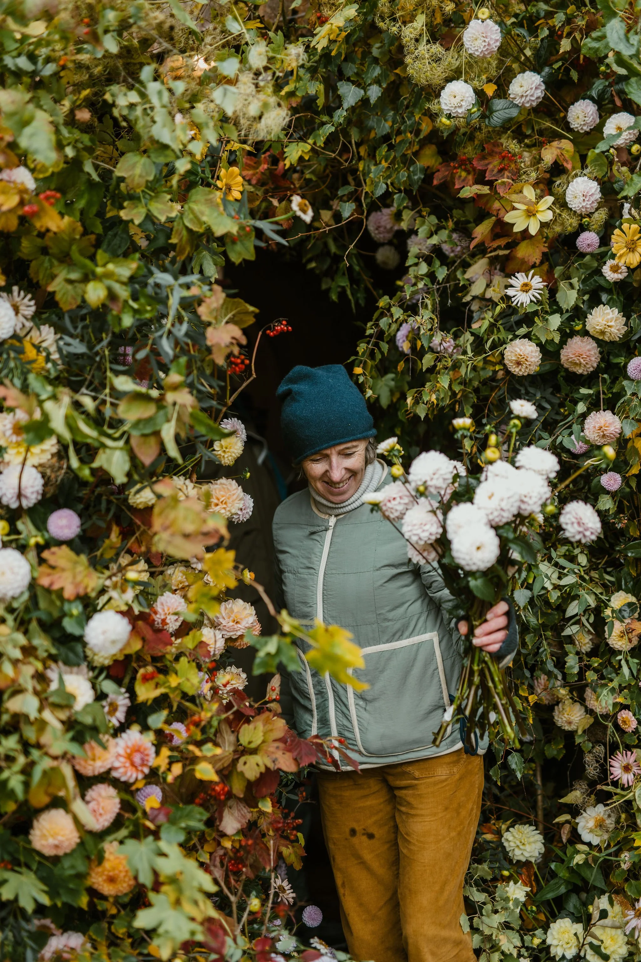 A woman wearing a dark beanie, light green jacket, and mustard-colored pants stands among a lush, colorful flower arch, smiling and holding a bundle of flowers.