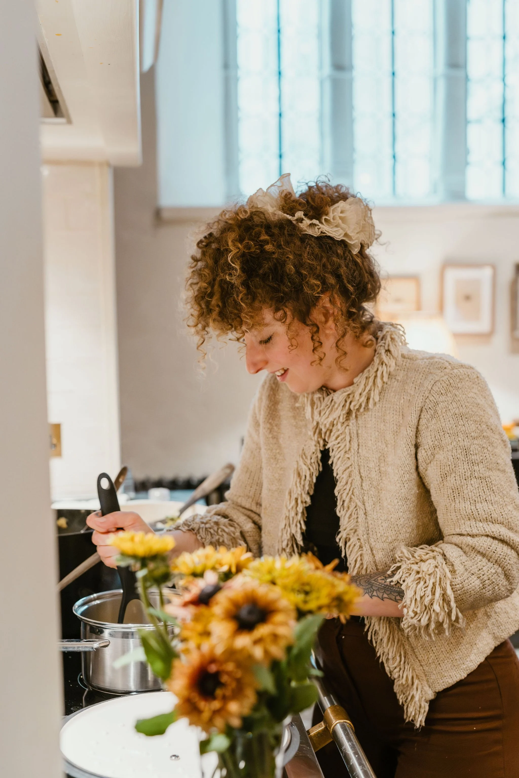 A woman with curly hair and a floral hair accessory cooking in a kitchen, smiling and preparing food surrounded by flowers and kitchen utensils.