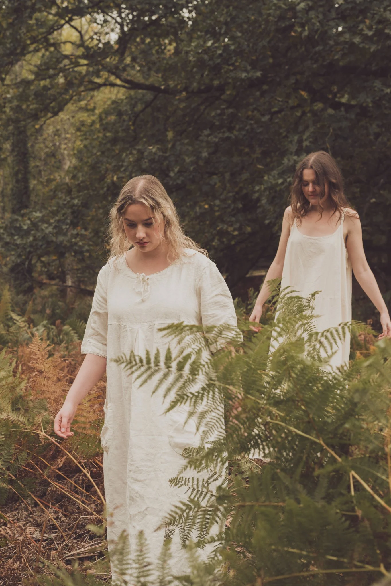 Two young women in cream-colored dresses walking through a forested area with ferns and trees.
