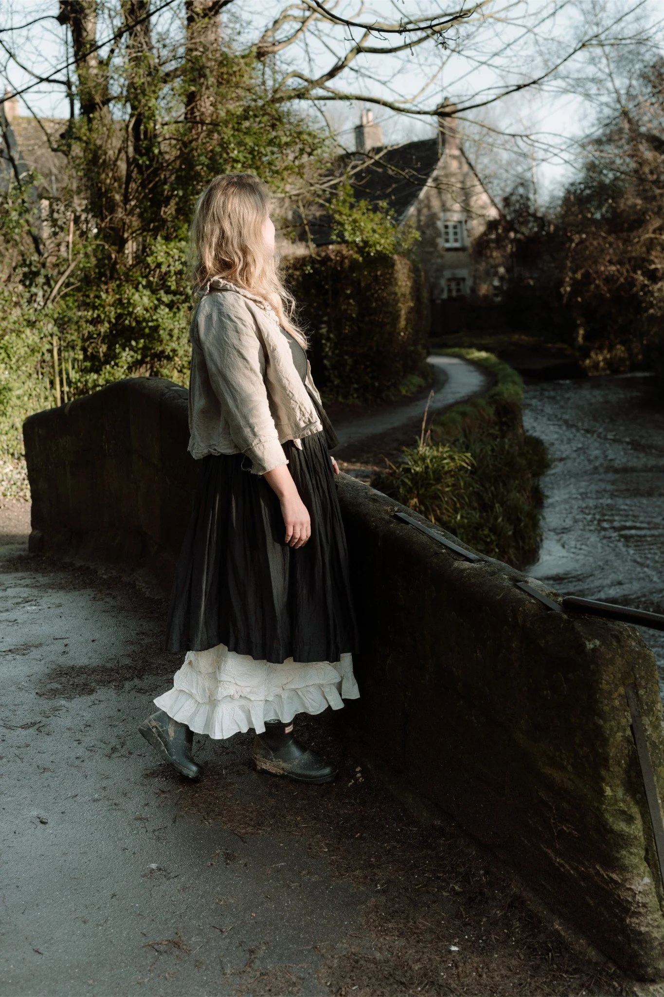 A woman wearing a long black and white dress with a beige jacket and black boots standing by a stone bridge overlooking a river in a park or garden area with trees and bushes in the background.