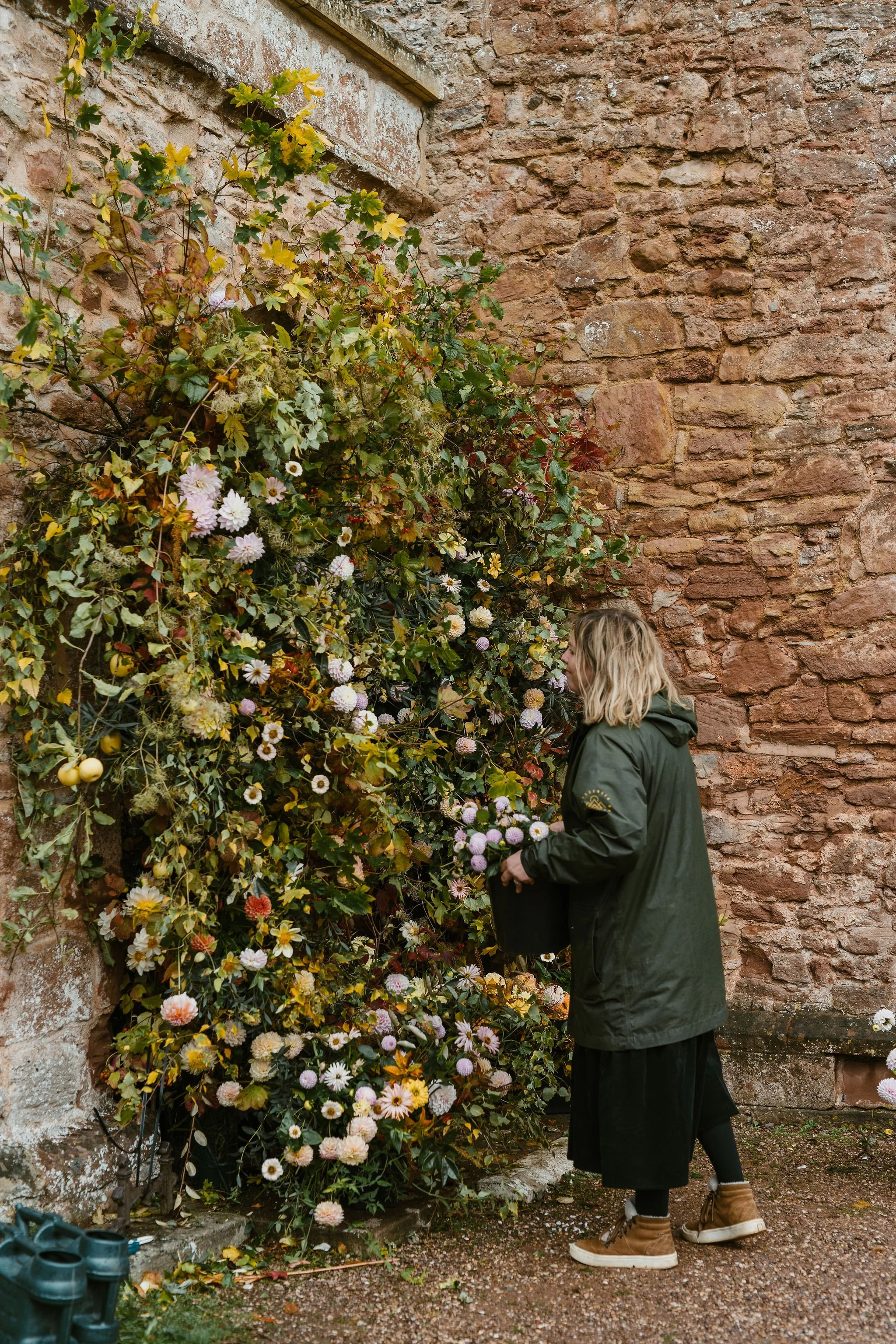 A woman wearing a black coat with blonde hair picking flowers from a large flower bush against a brick wall.