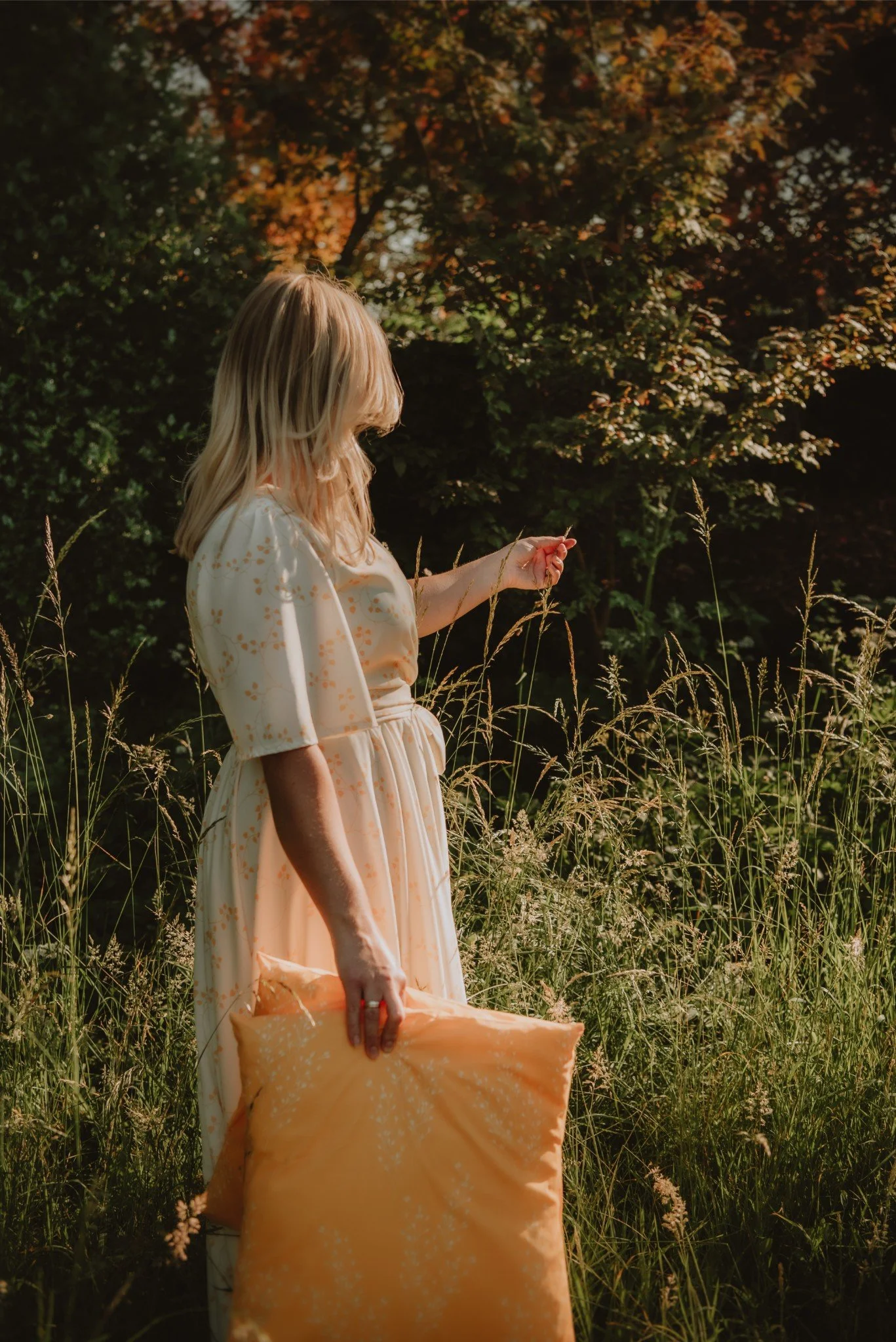 A woman with blonde hair wearing a light-colored dress standing in tall grass on a sunny day, holding a yellow pillow and a small flower, with trees and foliage in the background.