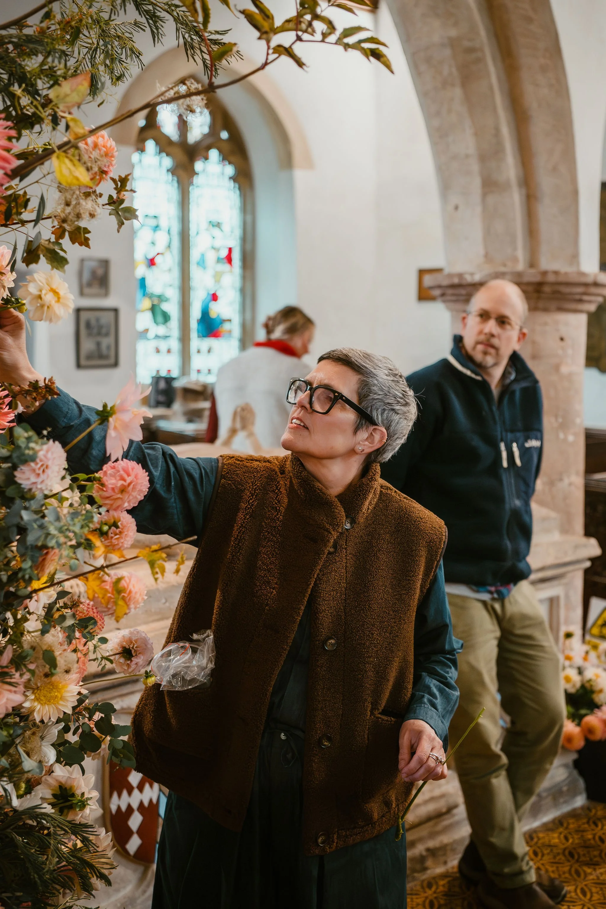 A woman in glasses and a brown vest arranging pink and cream flowers inside a church with stained glass windows, while two men observe.