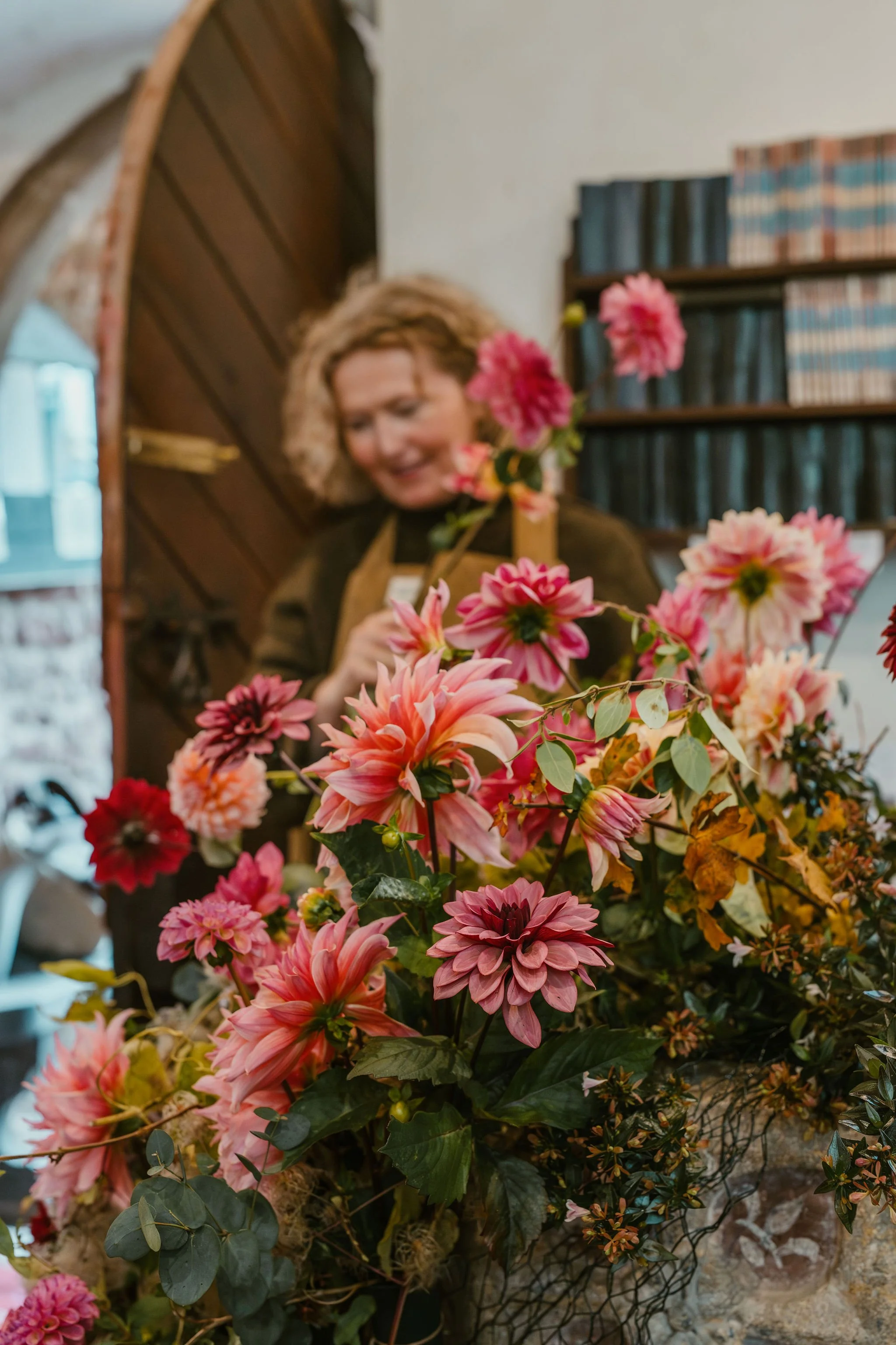 A woman arranging a large bouquet of pink and red dahlias with green leaves, in an indoor setting with bookshelf in the background.
