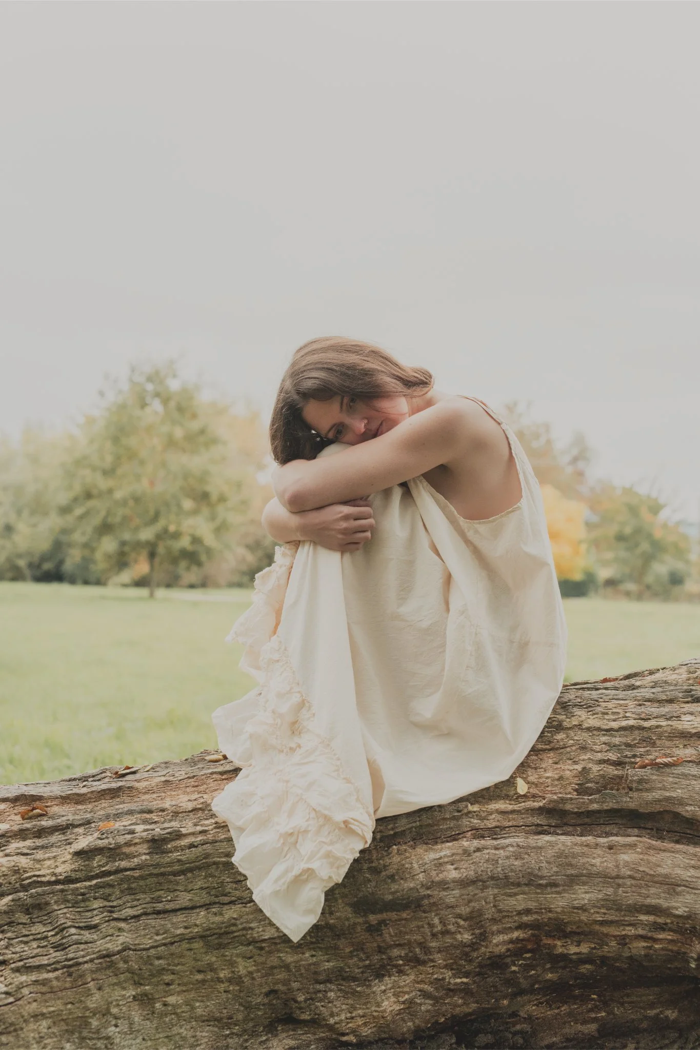 A woman with brown hair in a cream dress sits on a large log outdoors, hugging her knees, with a background of green grass and trees with fall foliage.