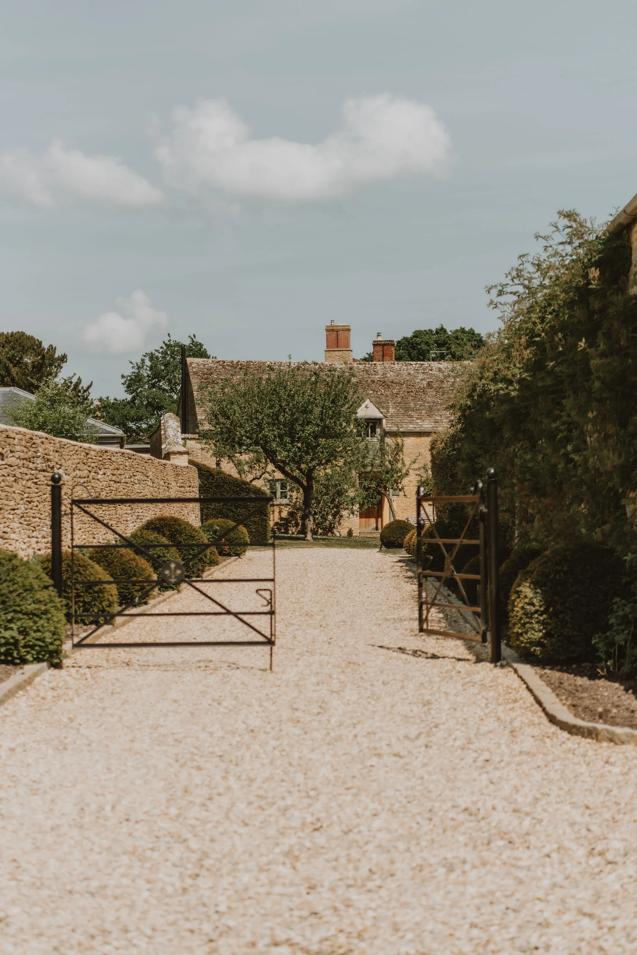 A gravel driveway with black metal gates opens to a courtyard surrounded by a stone wall and lush greenery, leading up to a stone house with chimneys under a partly cloudy sky.