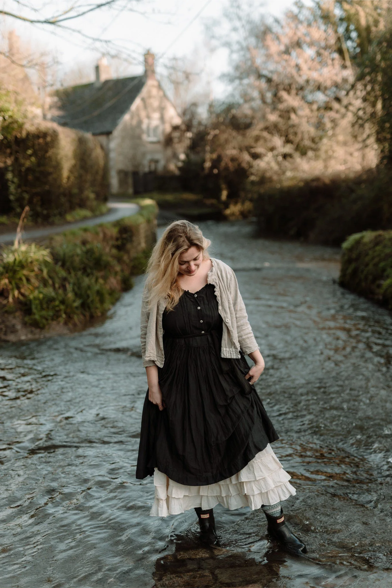 A woman standing in a shallow river with a dark dress, layered with a cream-colored skirt underneath, wearing black boots, and a light jacket, looking down with a smile in a rural setting with trees, a pathway, and a house in the background.