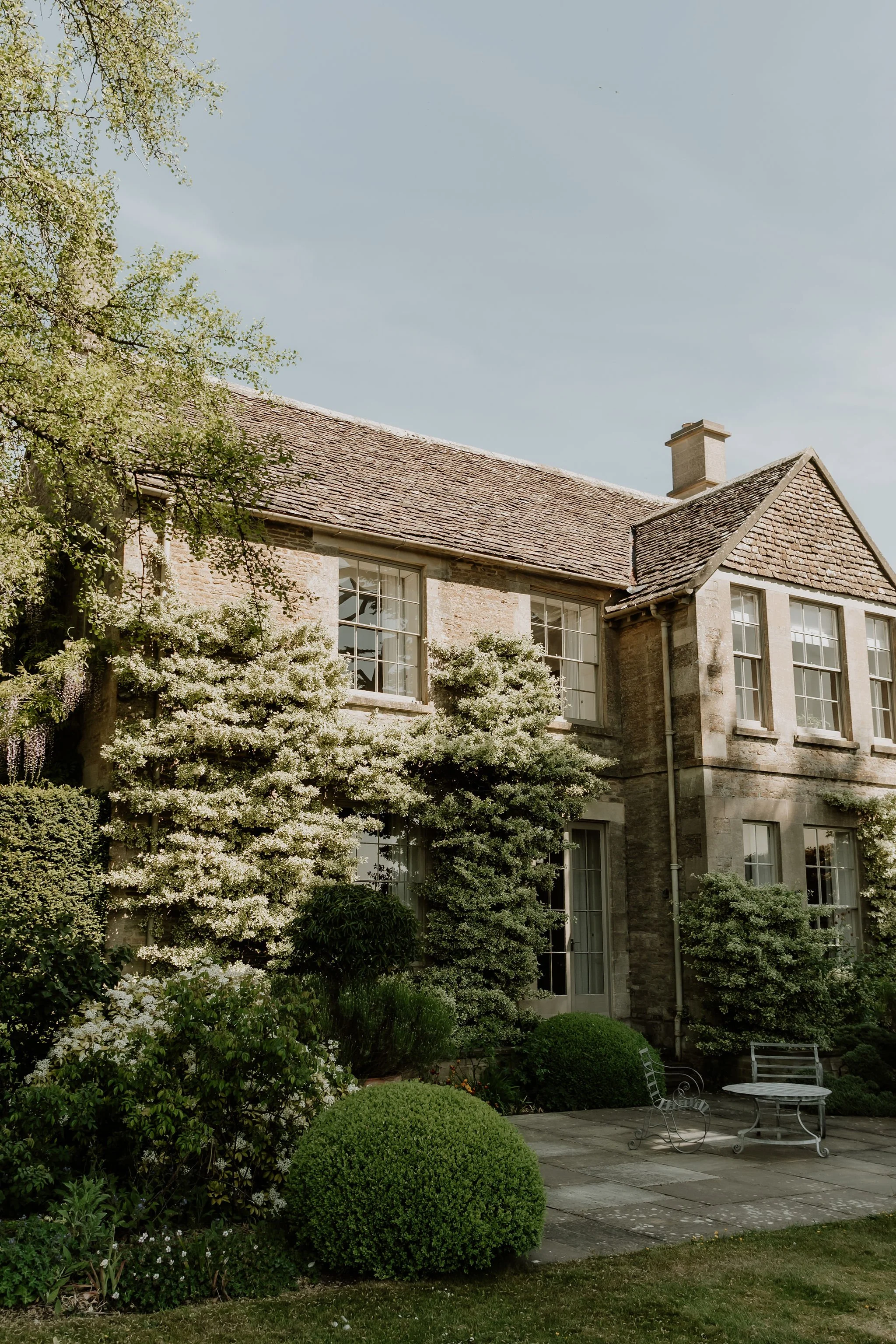 A two-story brick house with large windows, surrounded by lush greenery and flowering bushes, and a stone-paved patio with outdoor furniture.