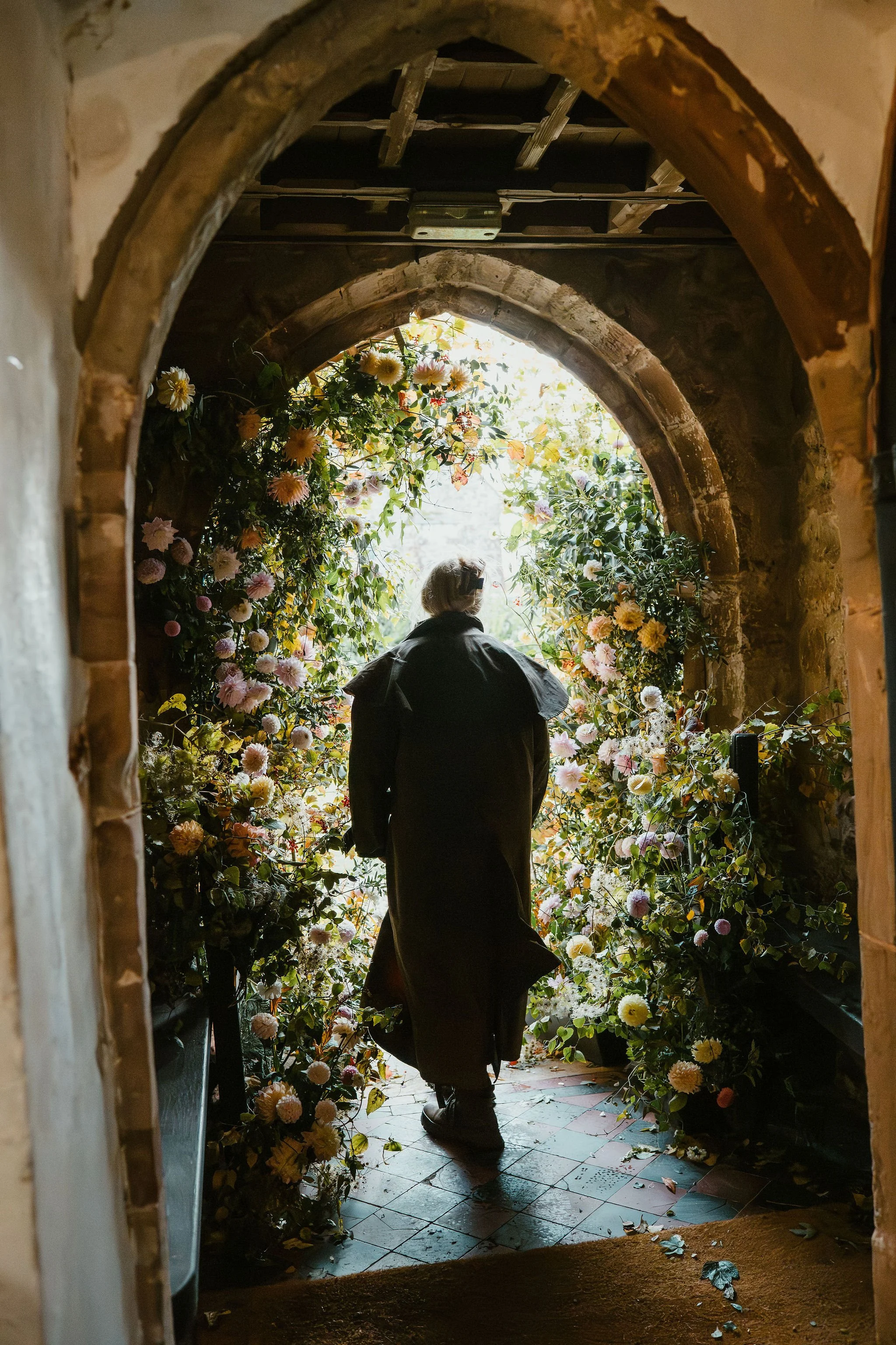 Person walking through an arched wood doorway surrounded by lush colorful flowers.