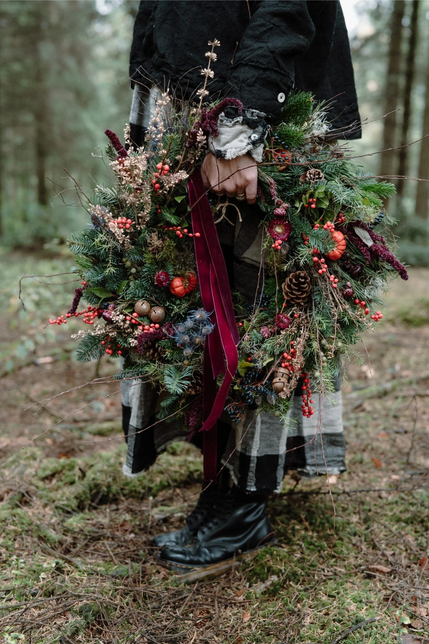 Person holding a Christmas wreath made of pine branches, berries, and small pumpkins in a forest setting.