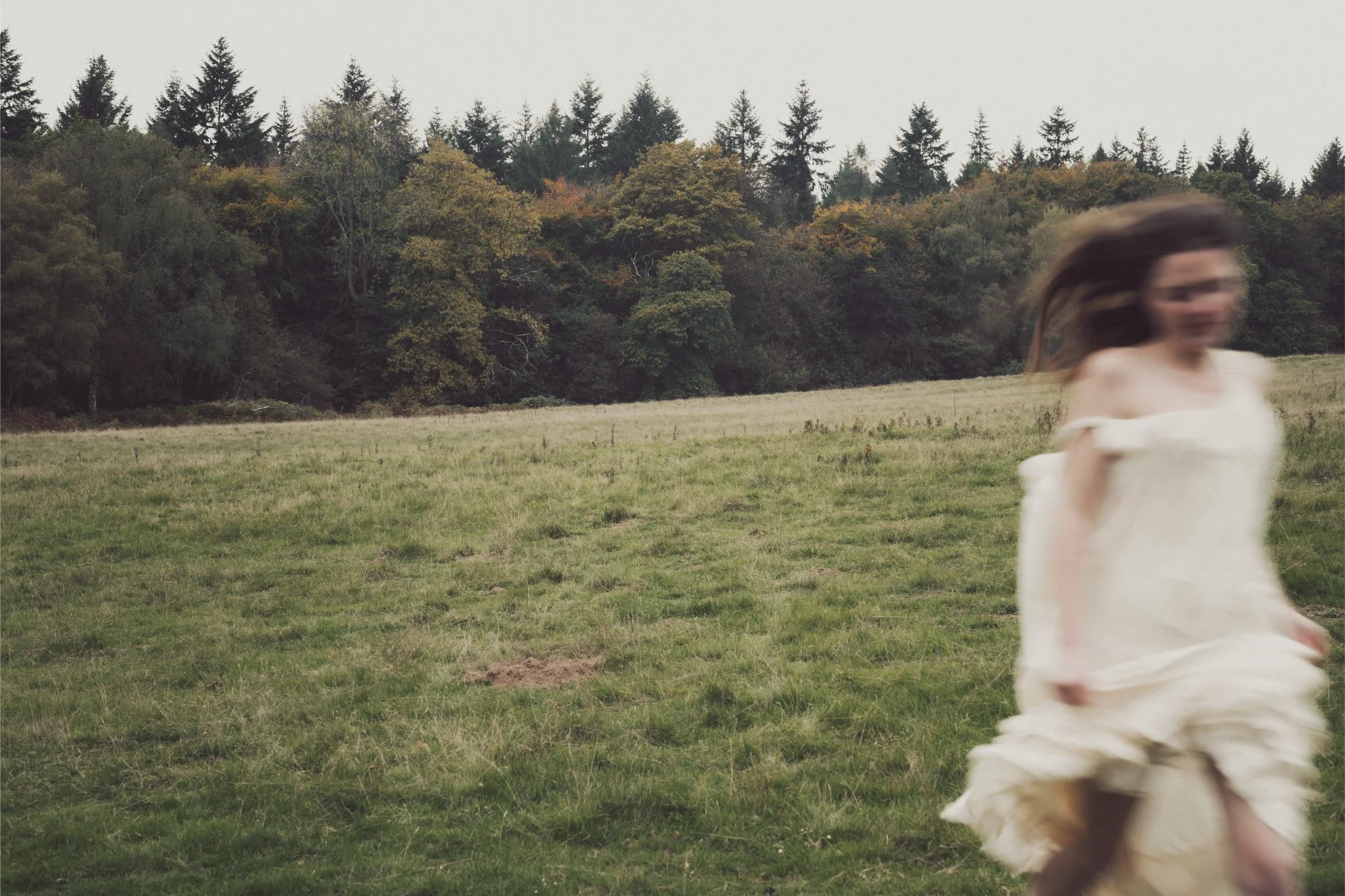A woman with long hair wearing a white off-shoulder dress walking across an open grassy field during daytime.