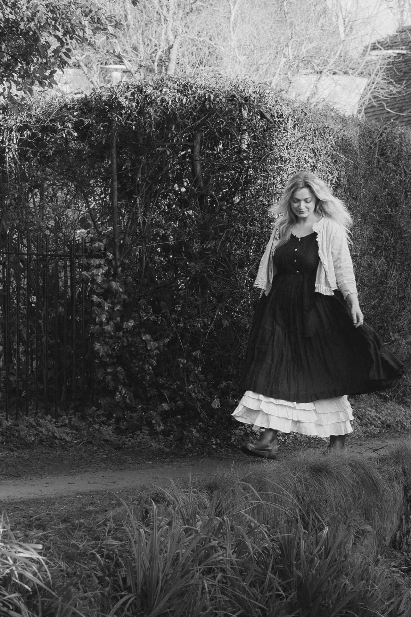 Woman walking along a garden path wearing a layered skirt and jacket, with trees and bushes in the background.