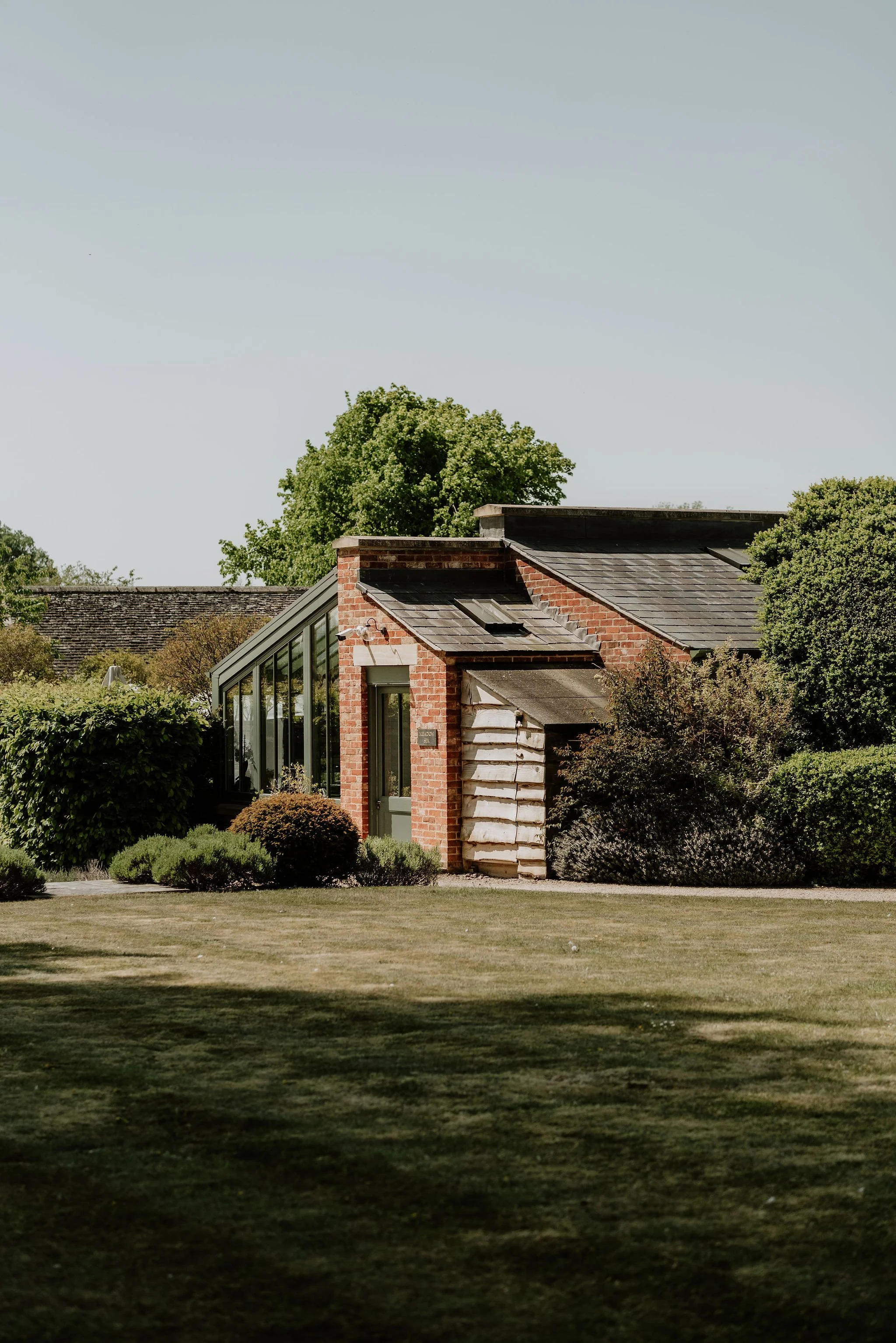 A small brick house with a modern glass extension, surrounded by well-maintained greenery and a large open lawn.