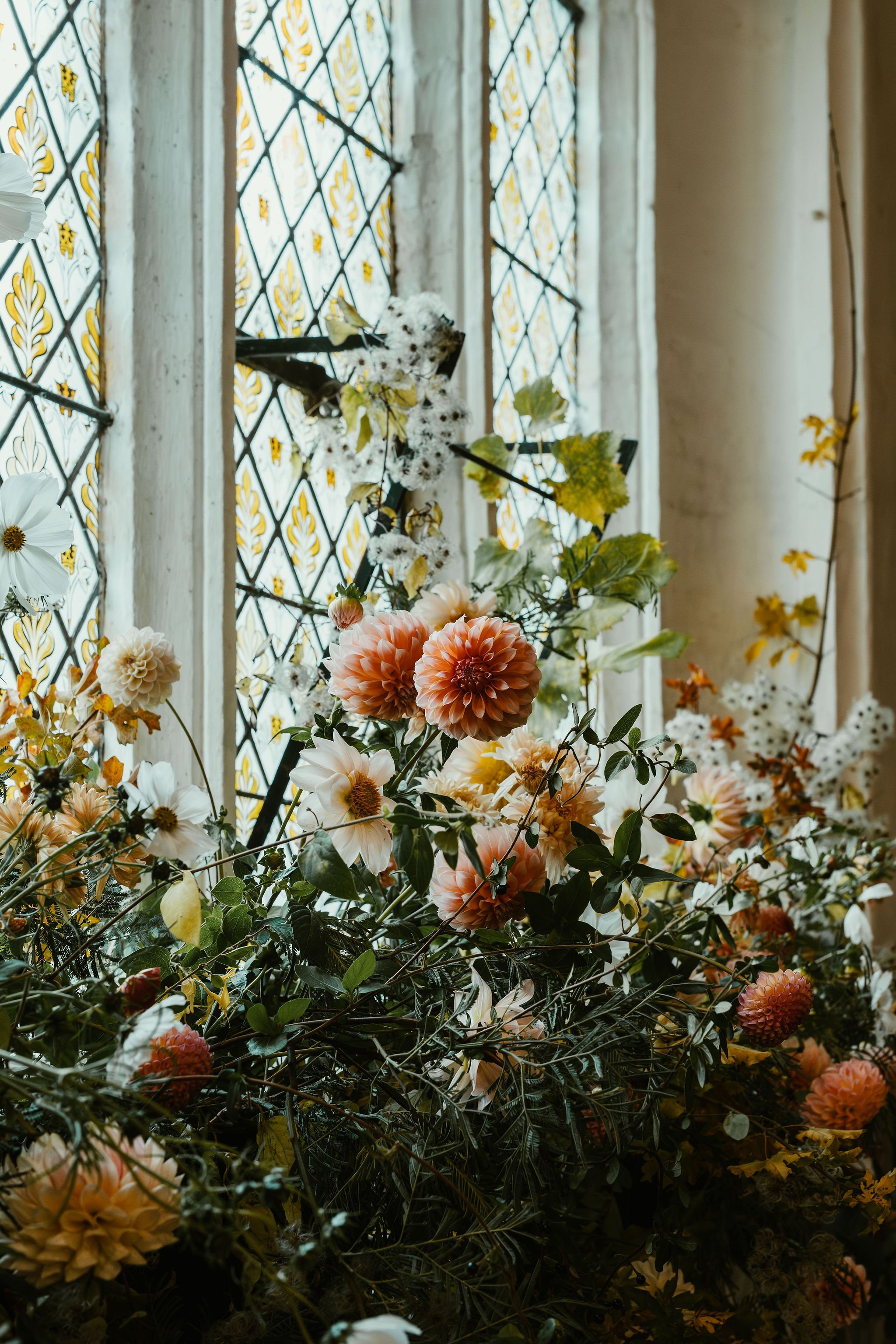 An indoor floral arrangement with pink, white, and peach flowers, green leaves, and a butterfly-shaped decoration, set near a stained glass window with yellow and black patterns.