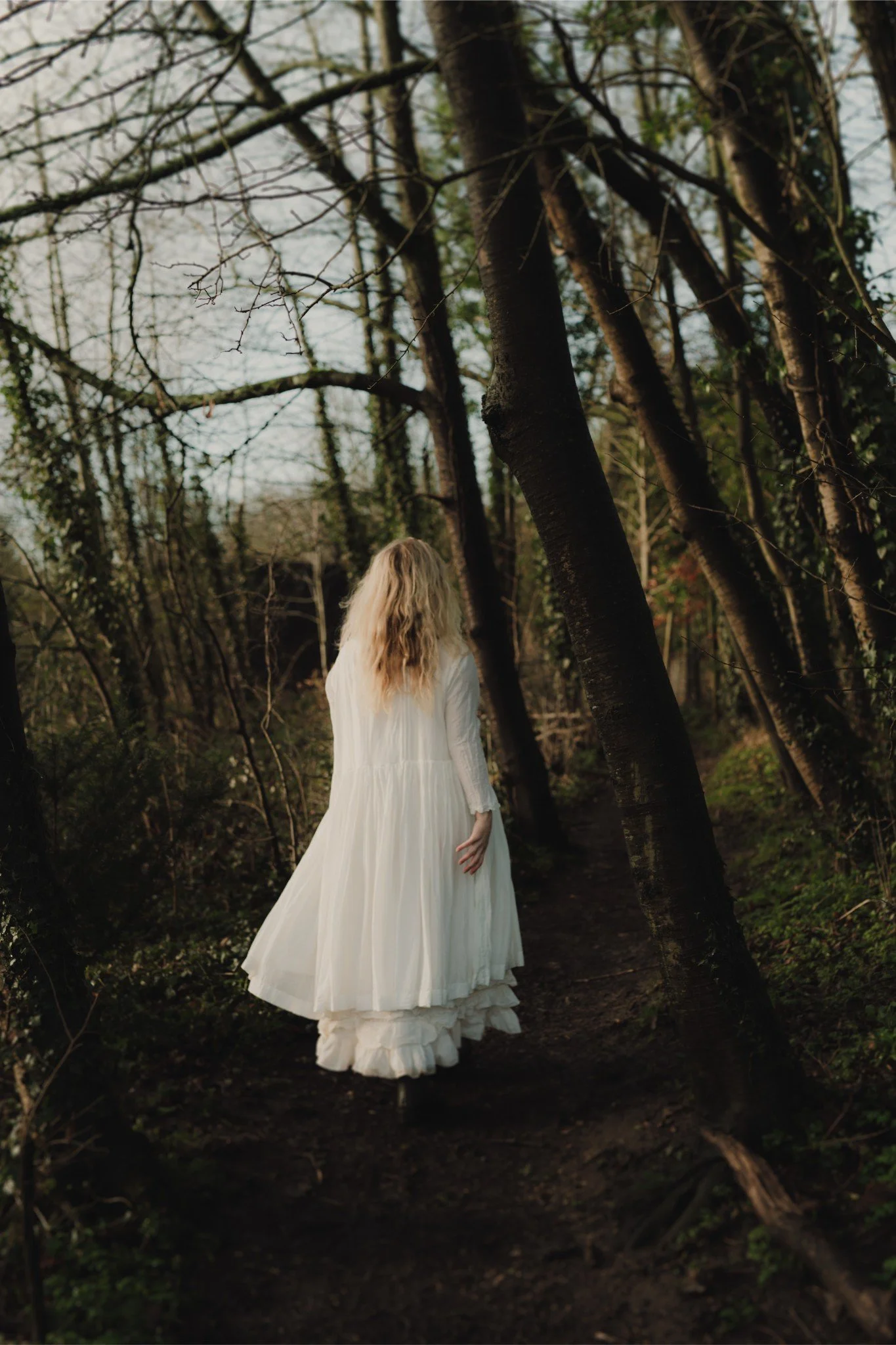 A woman with long blonde hair, dressed in a flowing white dress, walking along a dirt path through a wooded area with leafless trees.
