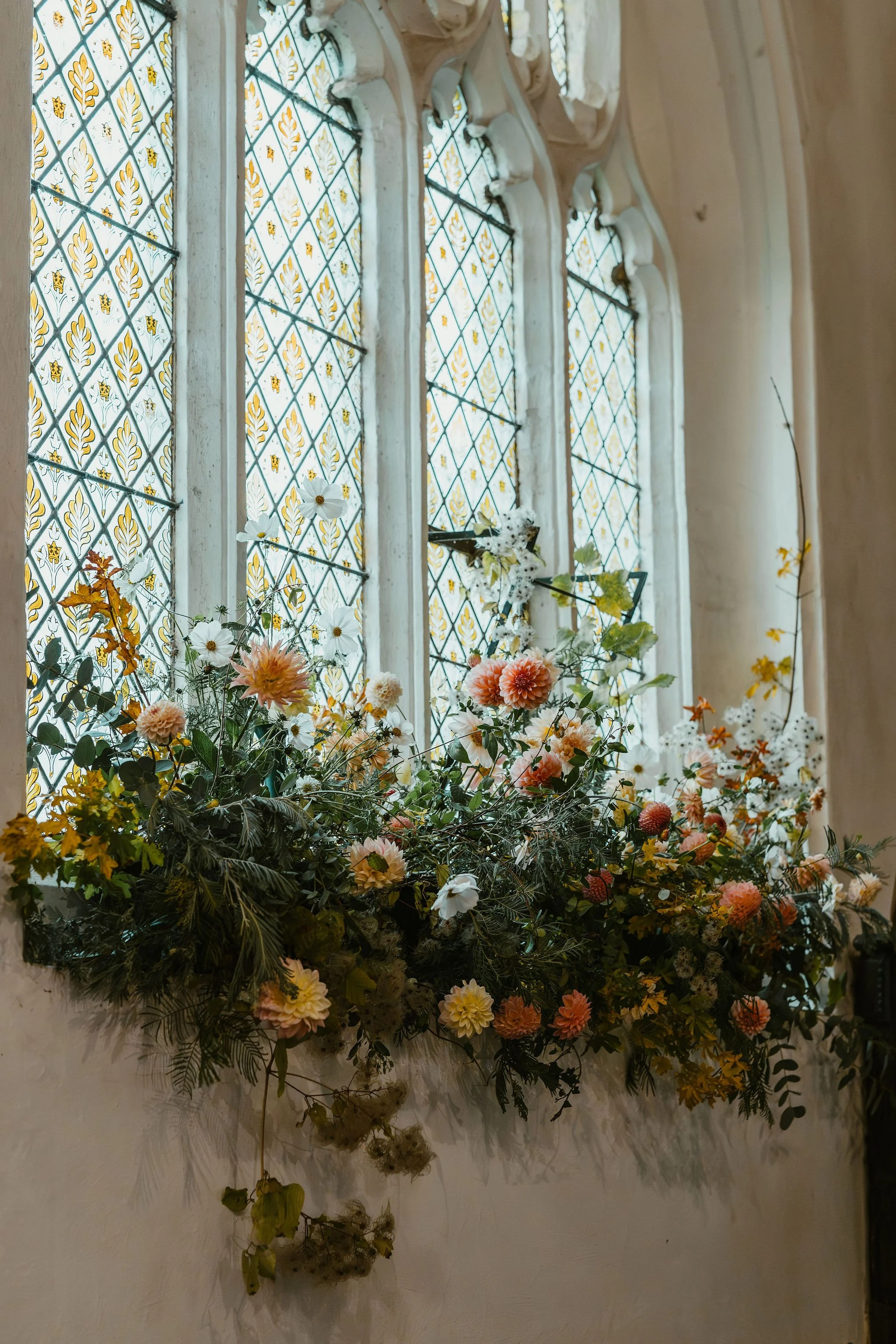 Colorful floral arrangement with pink, white, and yellow flowers on a windowsill inside a building with stained glass windows.