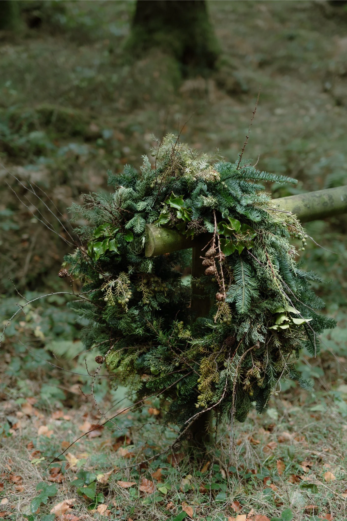 A Christmas wreath made of various evergreen branches and pine cones, hanging on a wooden post in a wooded area.