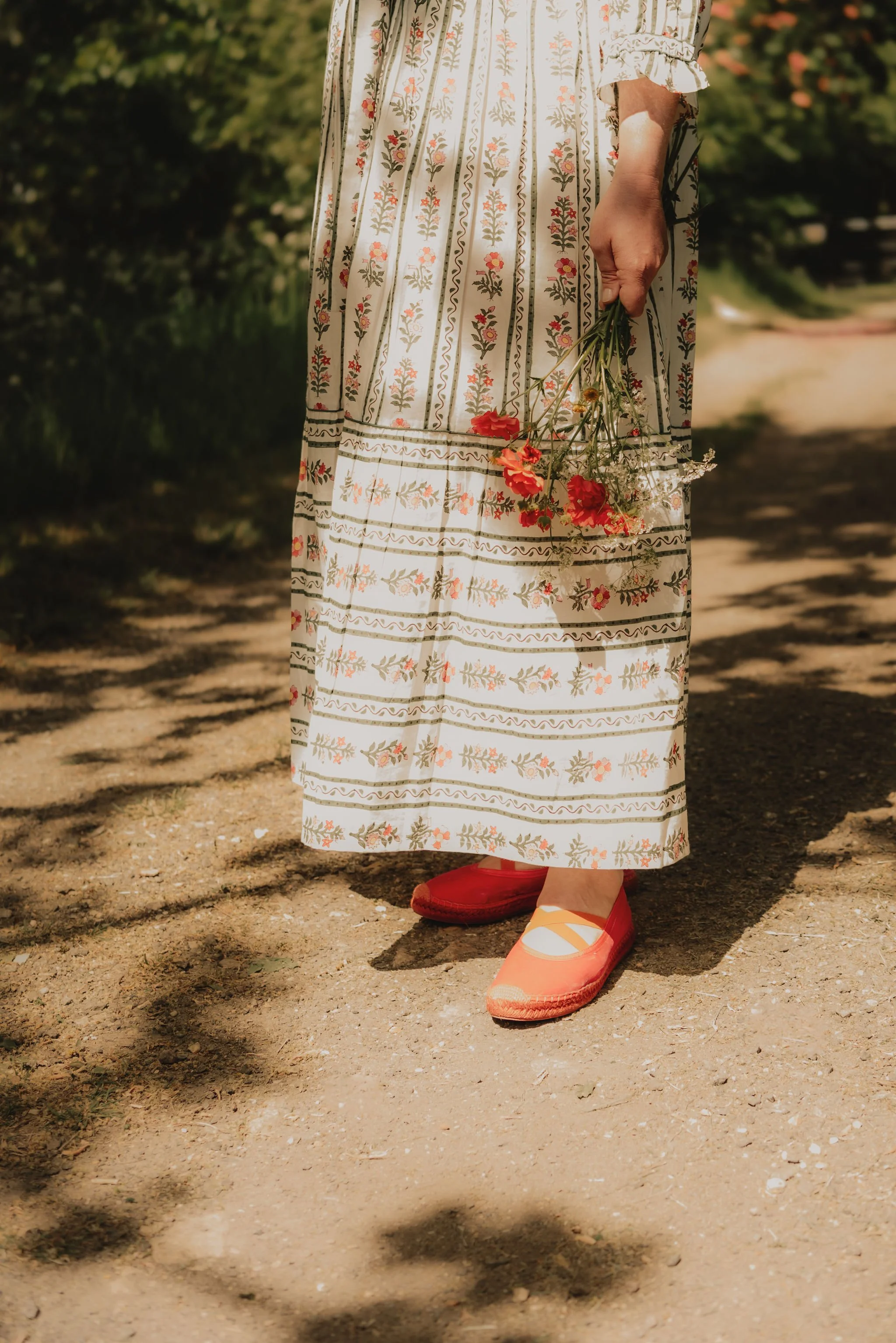 Person standing outdoors on a dirt path, wearing a long floral patterned dress and red shoes, holding a small bouquet of red and white flowers.