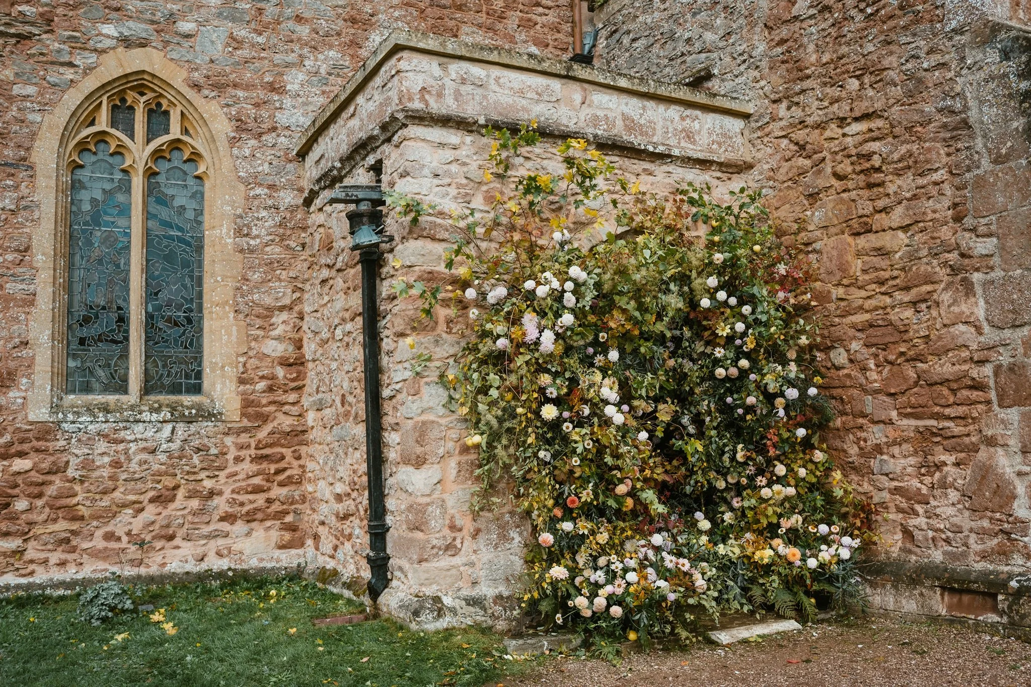 An exterior view of an old stone church wall with a stained glass window and a lush bush of white and pink flowers growing at the base.