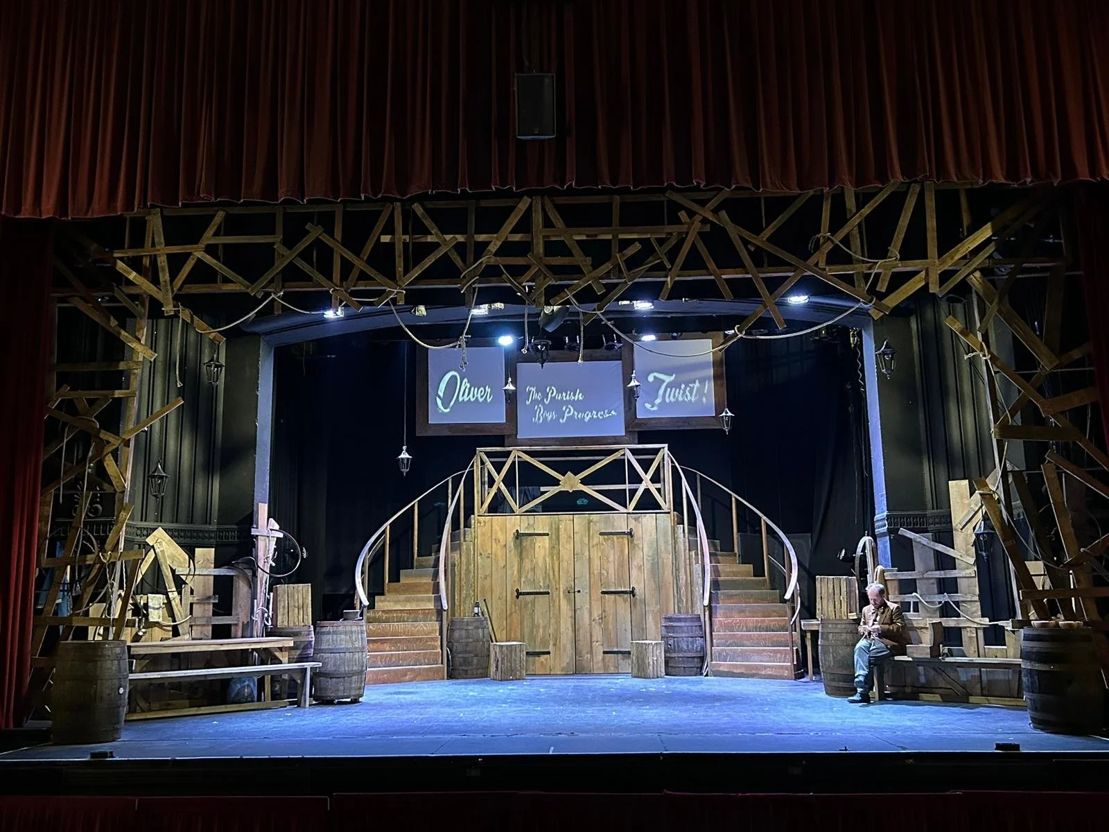 An image of the set of Oliver Twist at the Albany Theatre in Coventry.  Showing two curved wooden staircases centrally to the stage.  An actor is sat on scenery on the right hand side