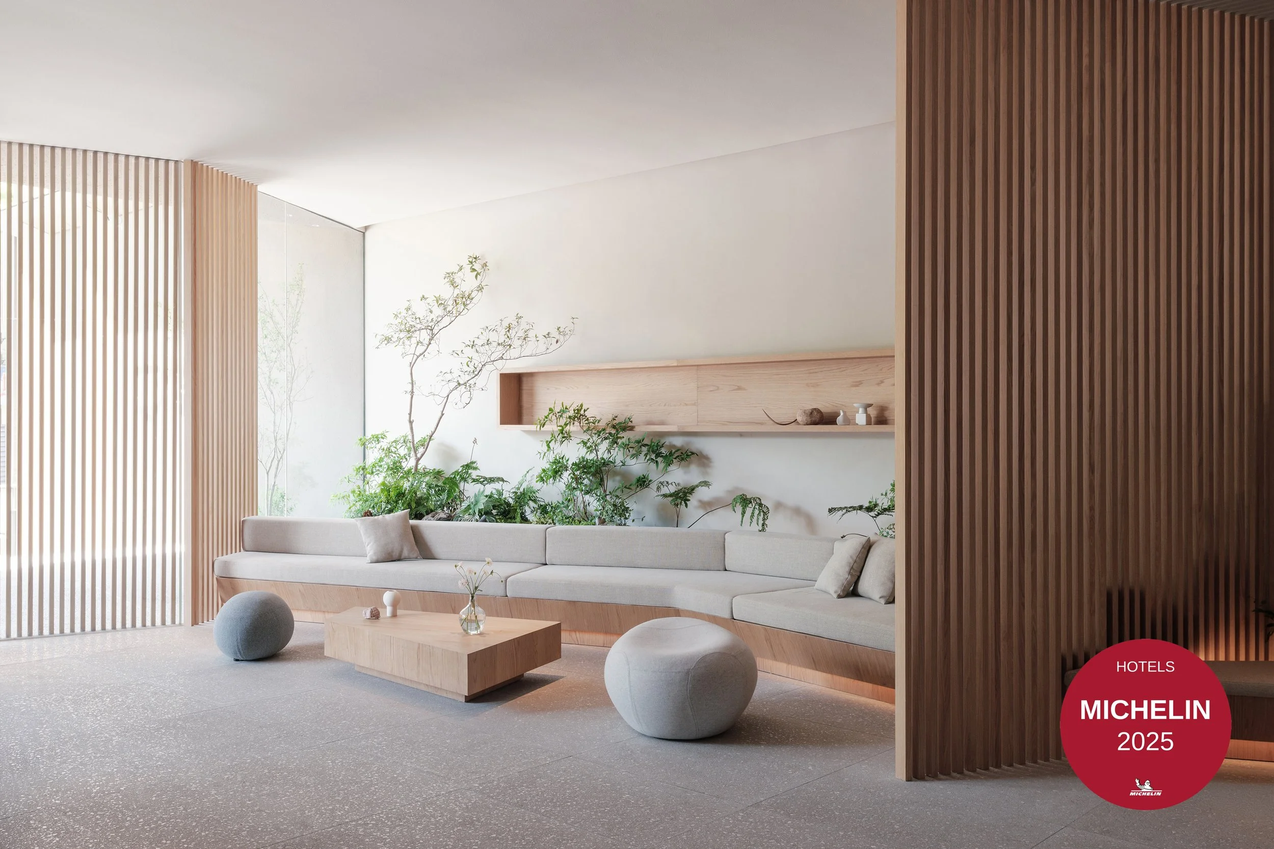 Modern living room with a beige sofa, wooden coffee table, and green indoor plants, separated by wooden slatted partitions, with natural light coming from large windows.