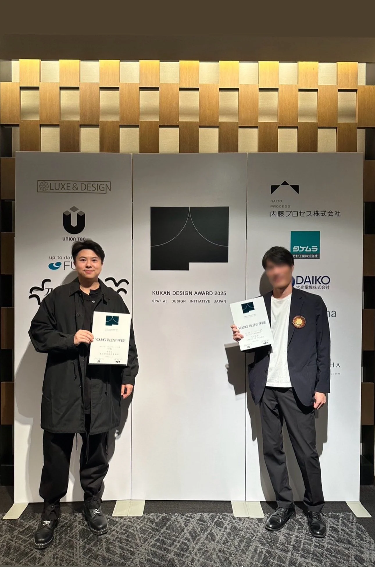 Two young men holding certificates at the Kukan Design Award 2025 event, standing in front of a white backdrop with logos and design graphics, in a well-lit indoor venue.