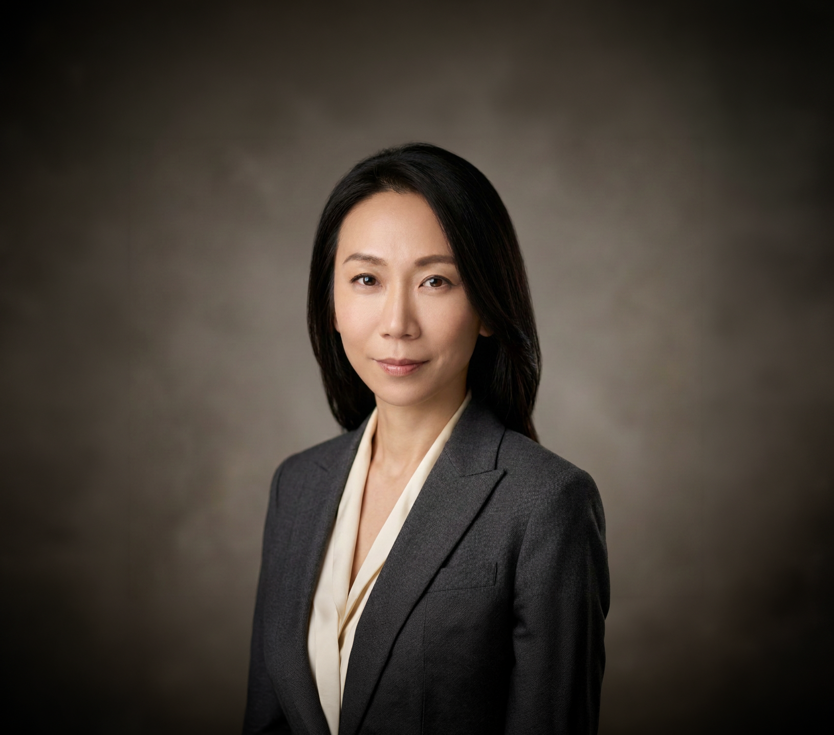 A professional woman with long dark hair wearing a gray blazer and white blouse, standing against a neutral background.