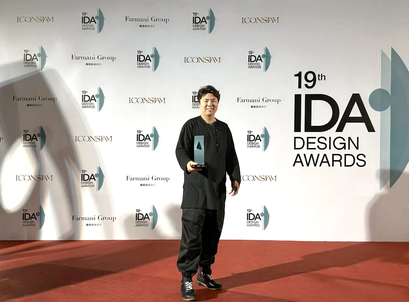 Person wearing black outfit holding a trophy at the 19th IDA Design Awards, standing in front of a backdrop with logos and text for the awards event.
