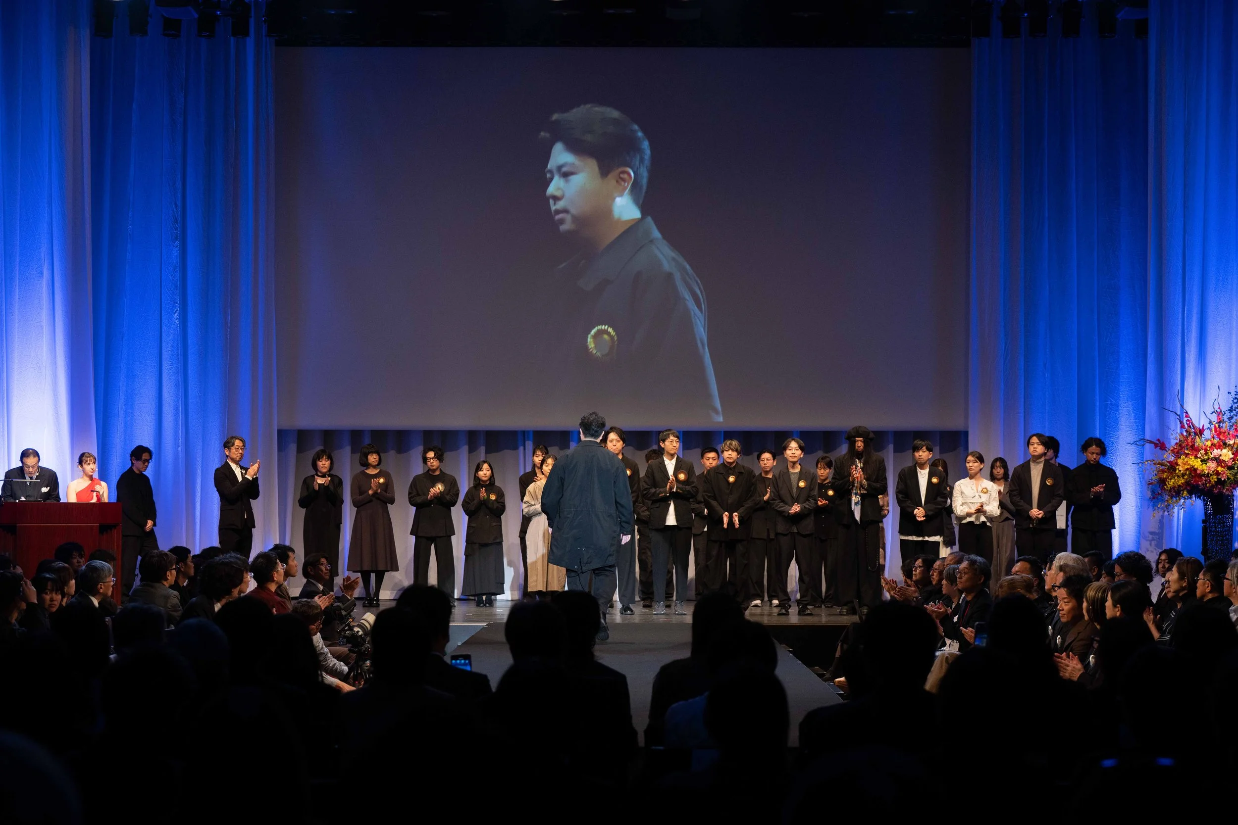 A large auditorium filled with people, with a stage where individuals are standing and clapping. A large screen displays a close-up image of a young man with dark hair in profile, wearing a dark jacket. The stage is decorated with blue curtains and colorful floral arrangements on the right side.