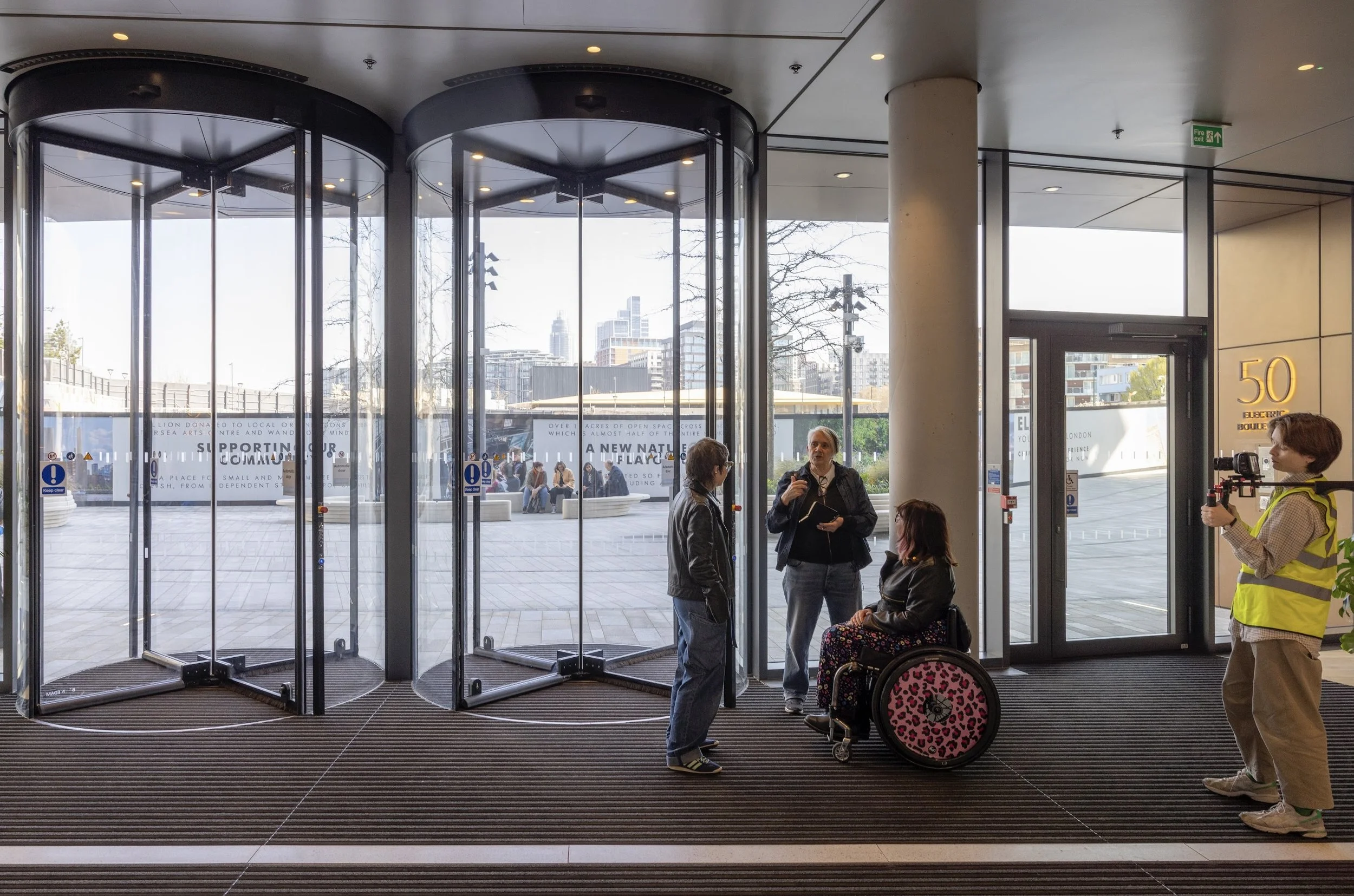 Photo of three people talking in front of a resolving lift. Two white women stand, one white woman with brown and pink hair sits in a wheelchair with pink patterned wheels.