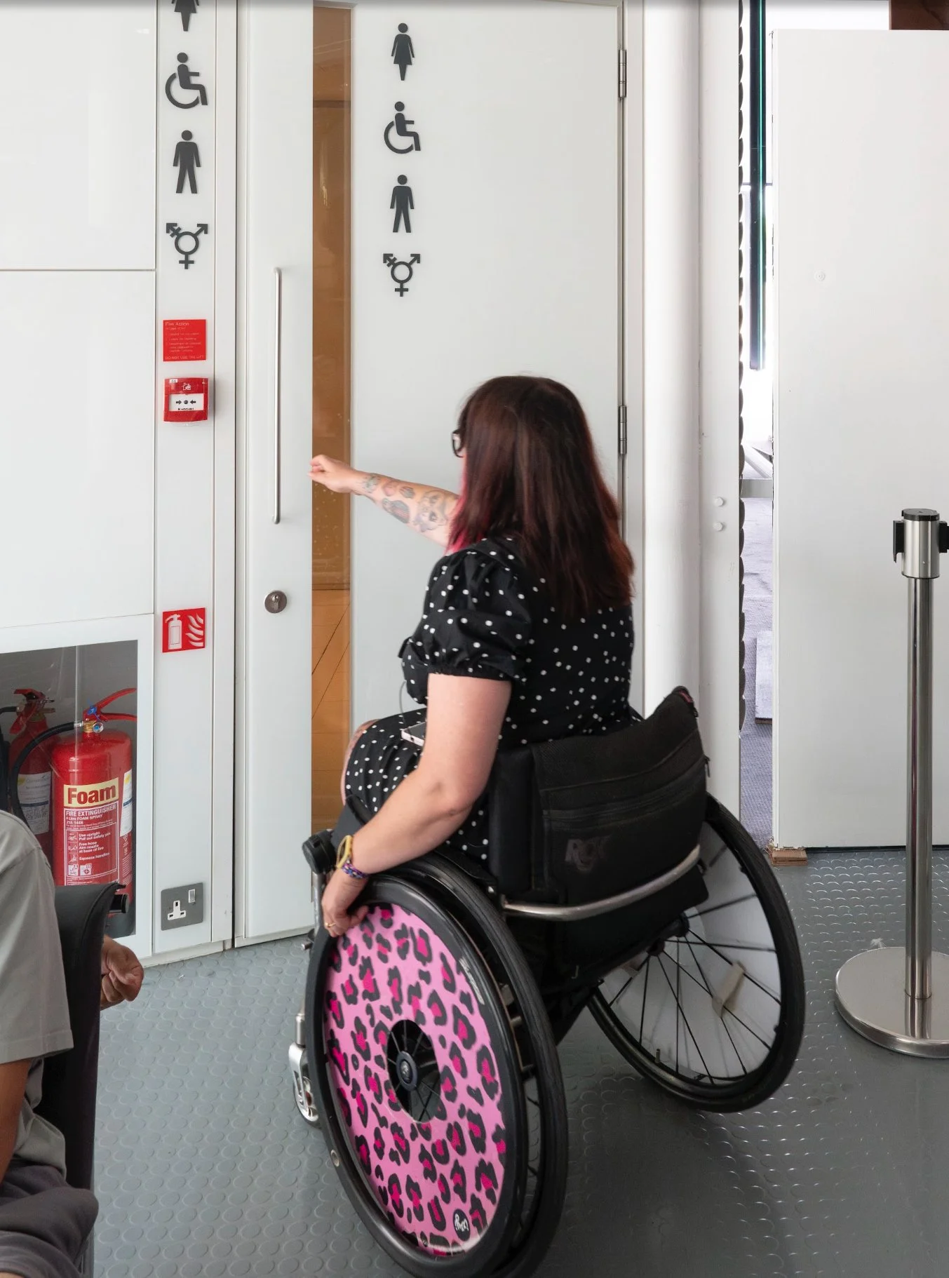 A woman in a wheelchair, with pink leopard print wheels, opens the door to the toilets in a white gallery space. 