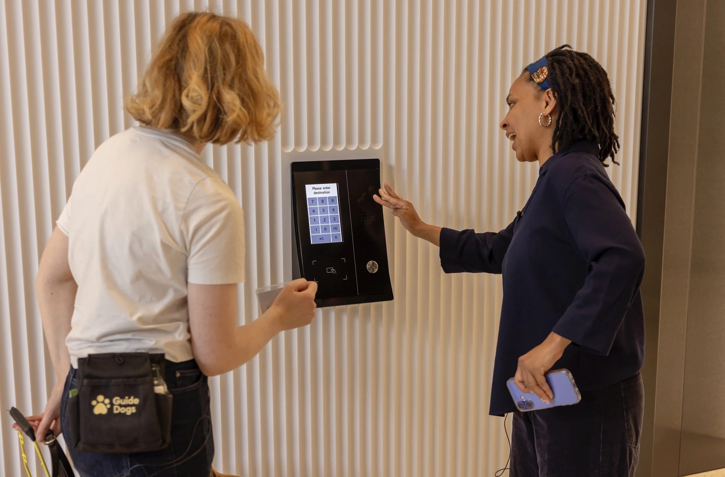 Photo of two women, looking at the buttons to a lift. One woman is wearing a guide dog pouch. 
