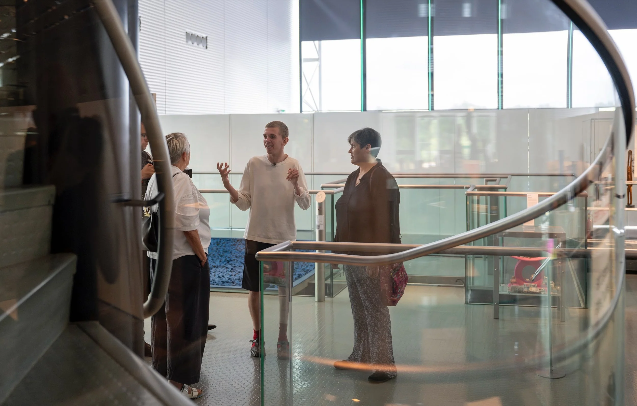 Three people talking, in a light filled gallery filled, with metal and glass railing around. 