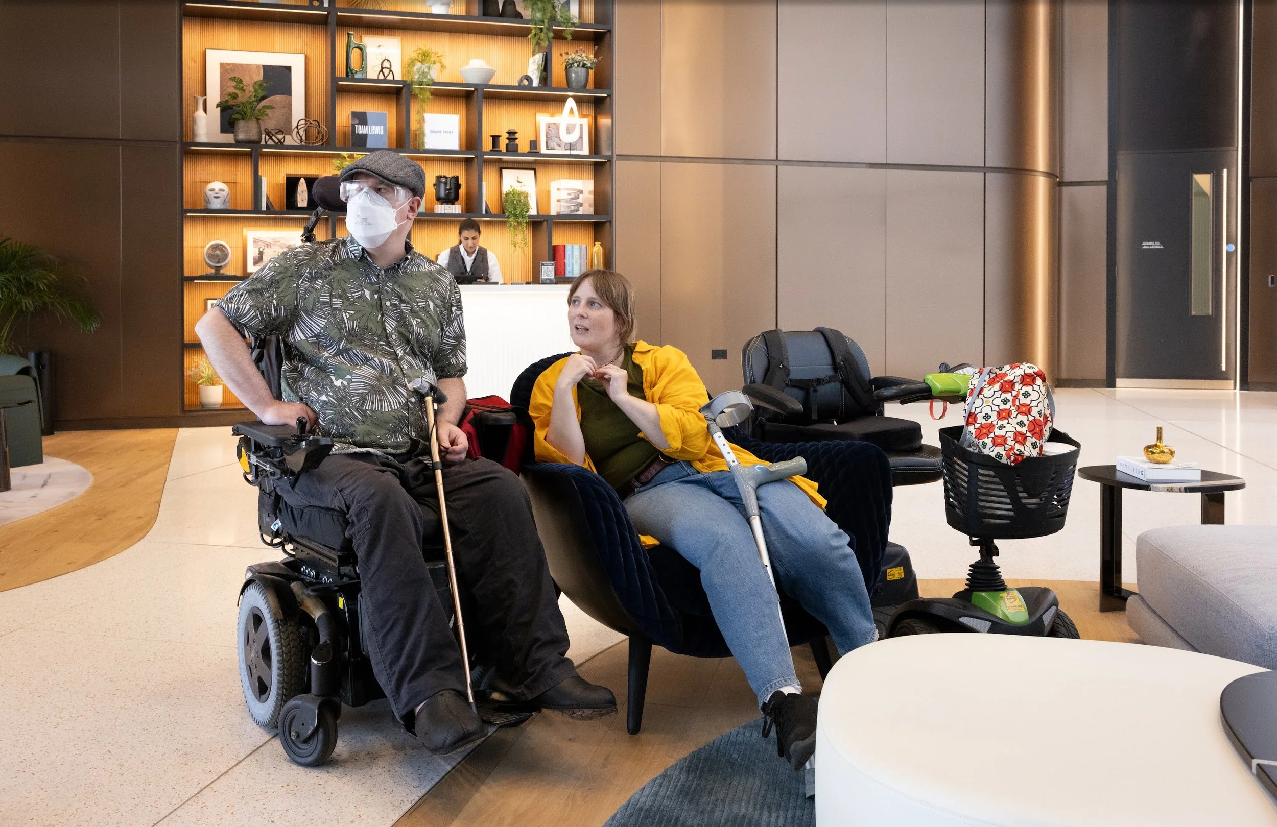 Two people talking in a modern lobby space, one man sat in a power chair with a mask and a woman sat in a comfy chair holding a crutch.