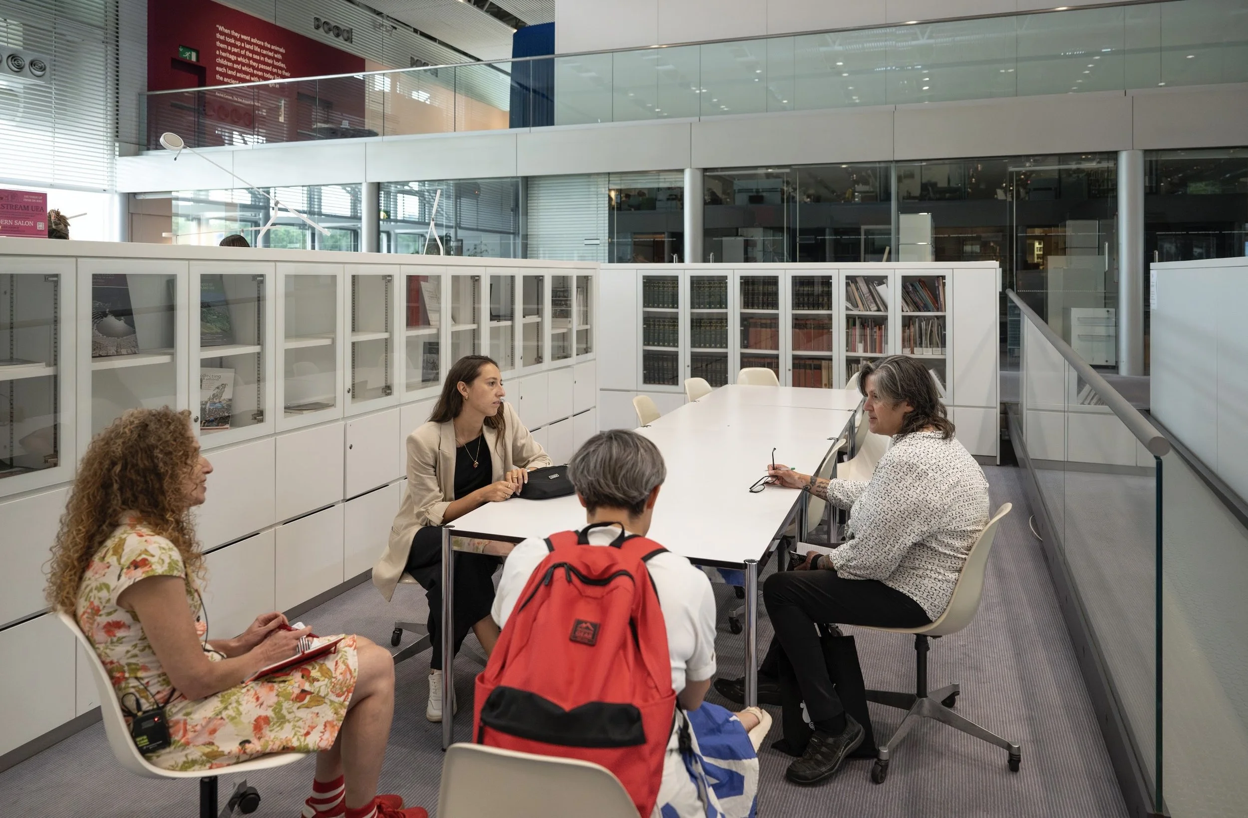 A group of four mixed women taking around a talking. They are in white study or library setting. 
