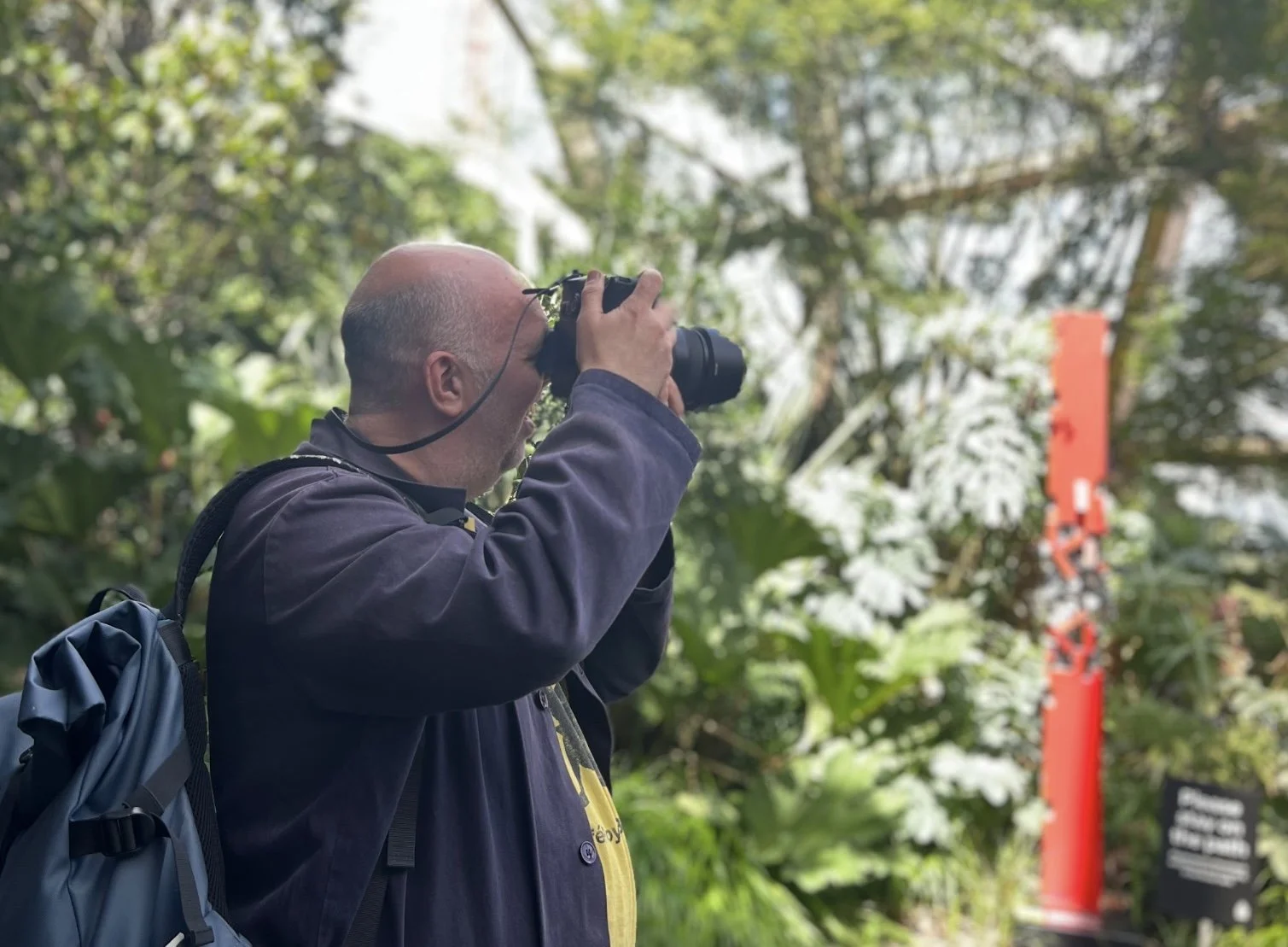 A white man wearing a dark blue jacket and rucksack taking a photograph of the surrounding in the Crossrail Place Roof Garden.