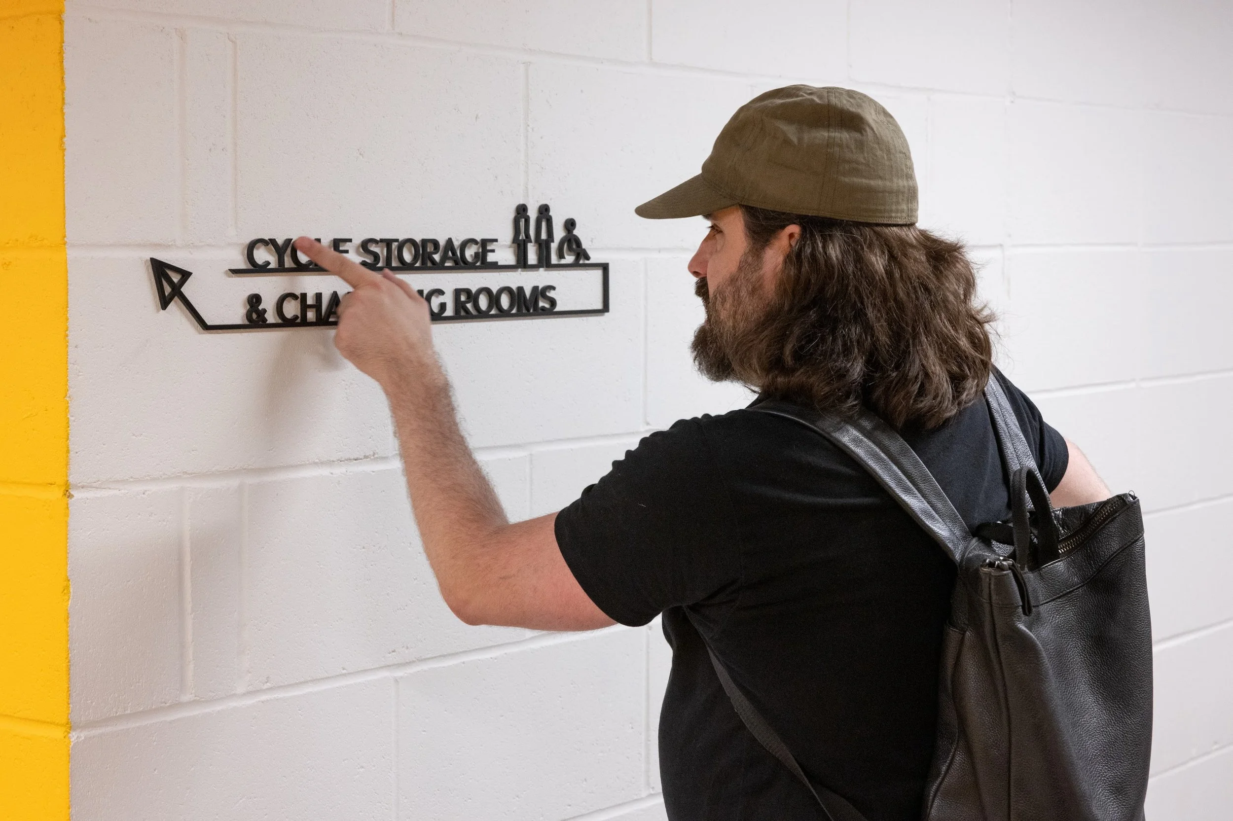 A man touching some 3D text on the wall, in black text on a white background, saying ‘cycle storage & changing rooms’ 
2. Two people talking in a modern lobby space, one man sat in a power chair with a mask and a woman sat in a comfy chair holding a 