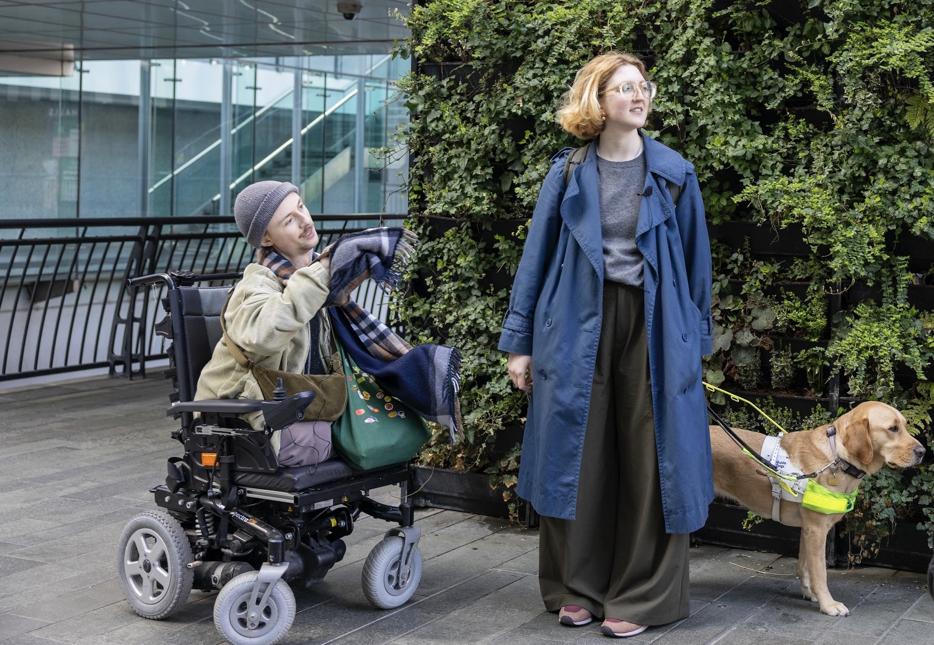 Photo of a white man in a power chair and a white woman with a guidedog look and point to the right, in front of a green wall and glasses building.