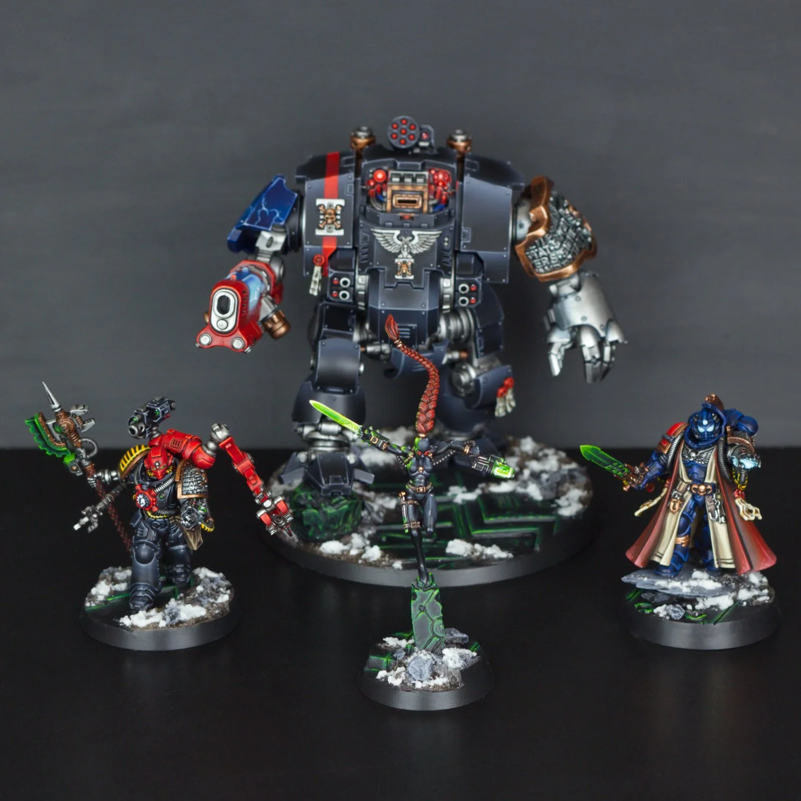 A collection of painted miniature figures from a tabletop game, depicting armored warriors in dynamic poses, with the central figure being a large mechanized robot with red and black armor, accompanied by smaller figures wielding swords and guns, all
